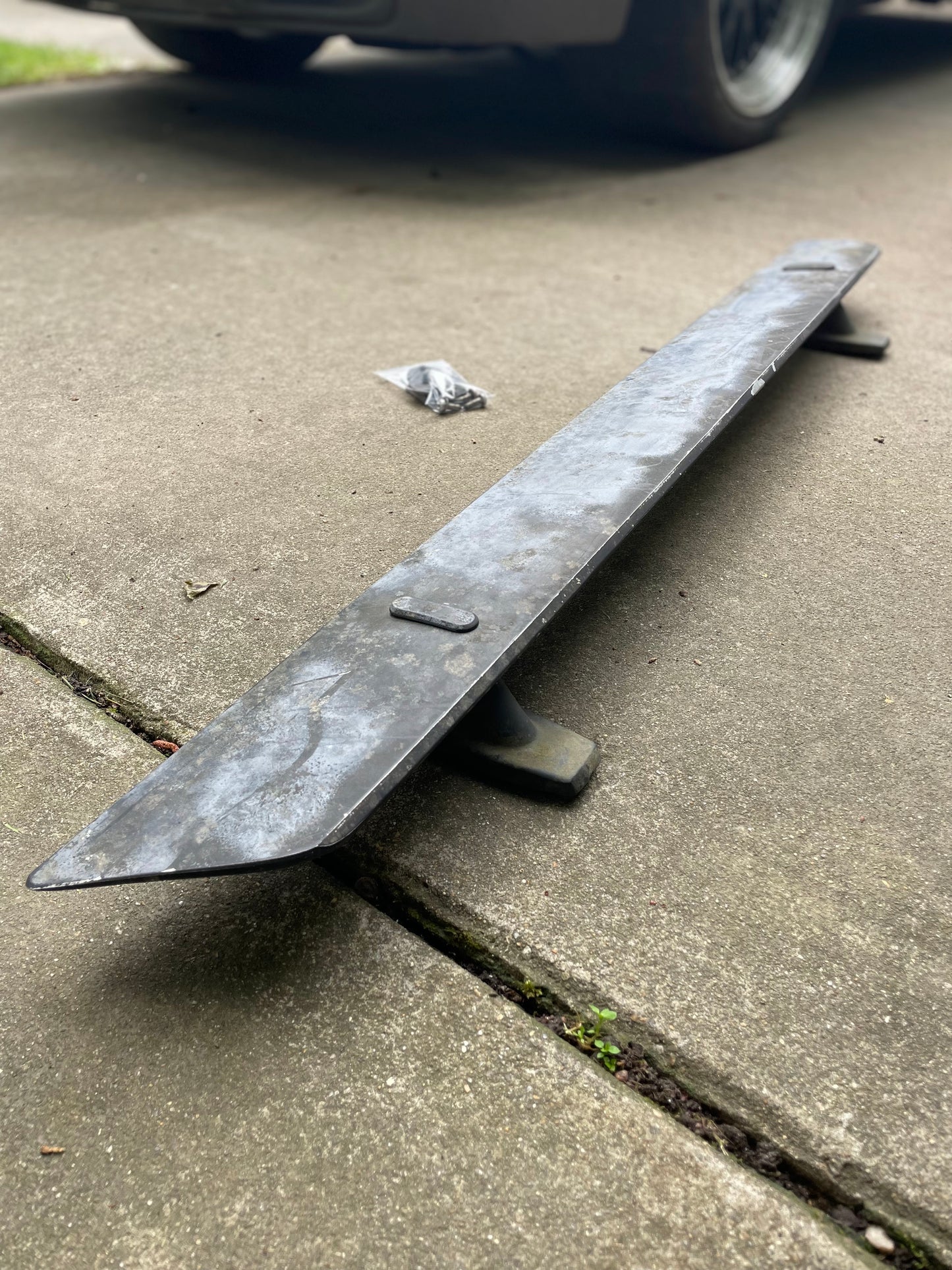 General Motors OEM Rear Hatch Hinges with Spoiler Wing for 1983-1994 Chevrolet S10 Blazer rest on a concrete driveway near a small bag of screws. Part of a car’s wheel and bumper appear in the background.