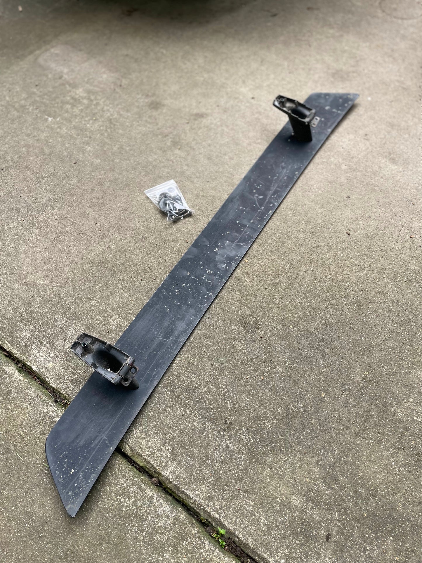 A General Motors OEM Rear Hatch Hinges with Spoiler Wing for 1983-1994 Chevrolet S10 Blazer is displayed on concrete, with a small bag holding screws and mounting hardware placed nearby.