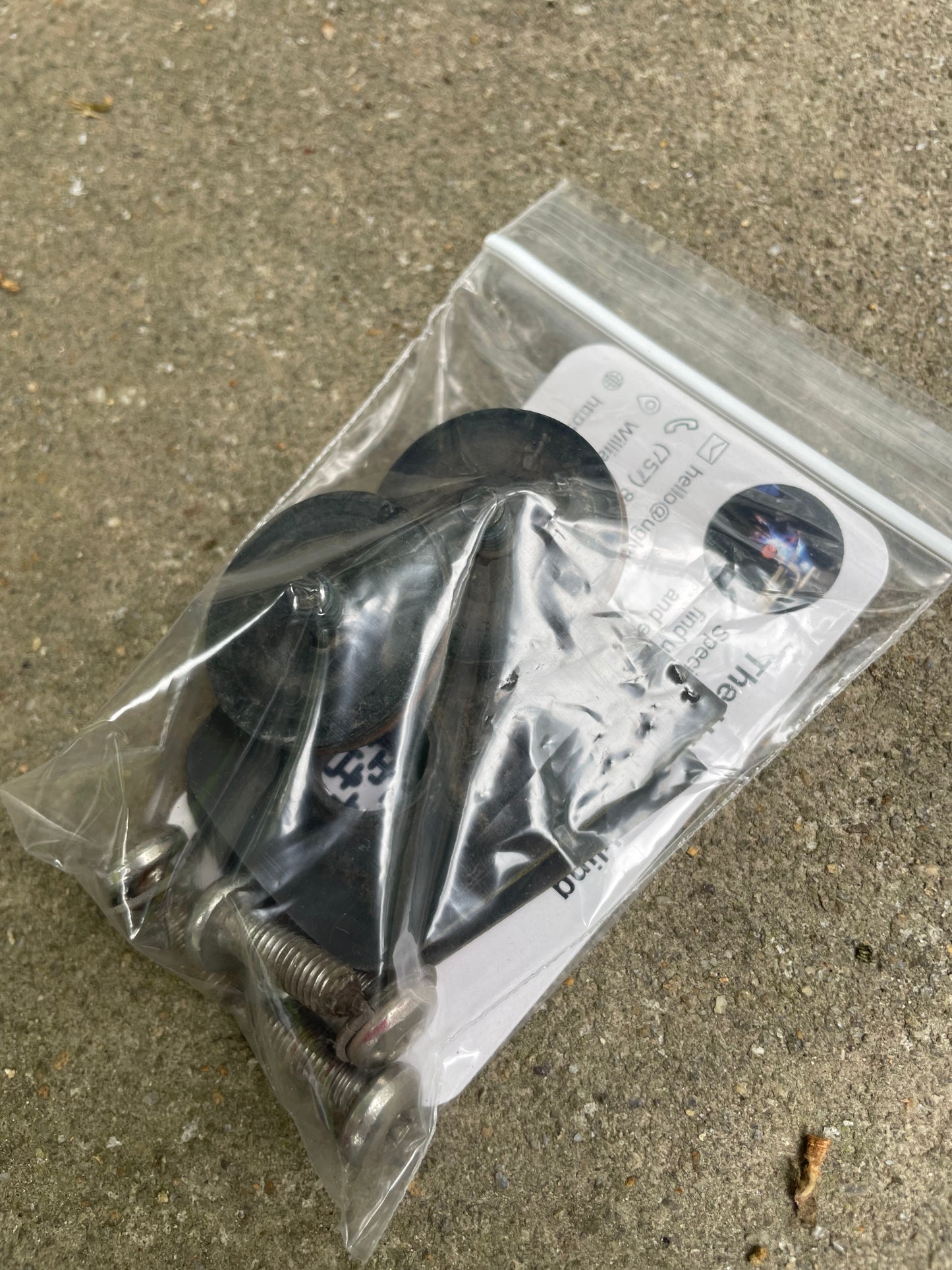 A clear bag with black rubber washers, metal screws, and an instruction sheet for the General Motors OEM Rear Hatch Hinges with Spoiler Wing (1983-1994 Chevrolet S10 Blazer and more) rests on rough concrete.