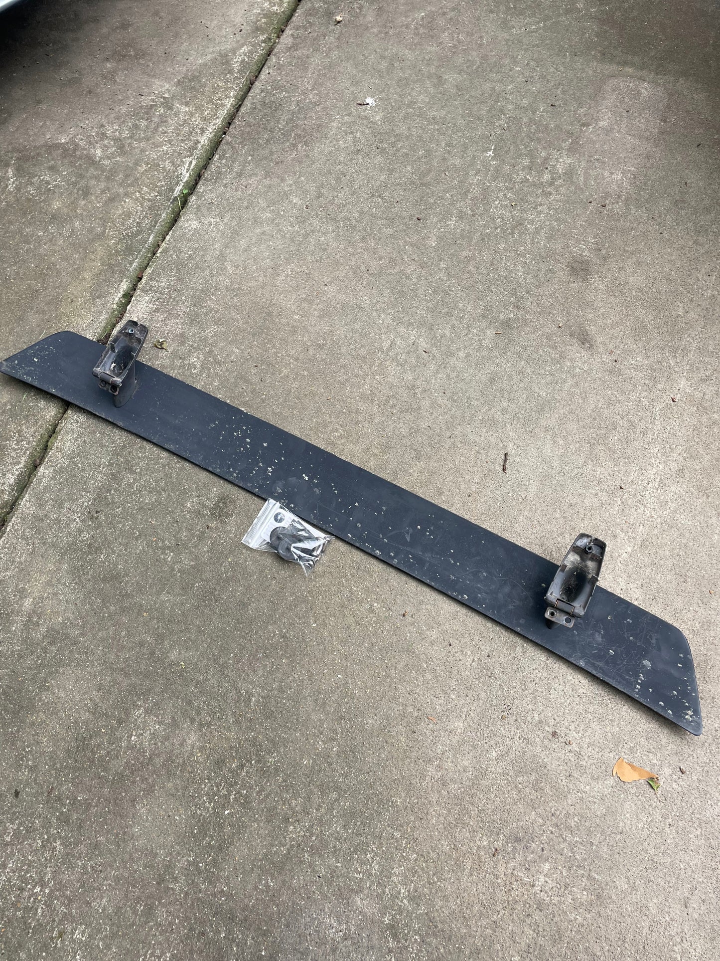 An OEM Rear Hatch Hinges with Spoiler Wing set by General Motors for 1983-1994 Chevrolet S10 Blazer and others, including two brackets and a small hardware bag, is shown on concrete. The metal parts appear dirty and weathered.
