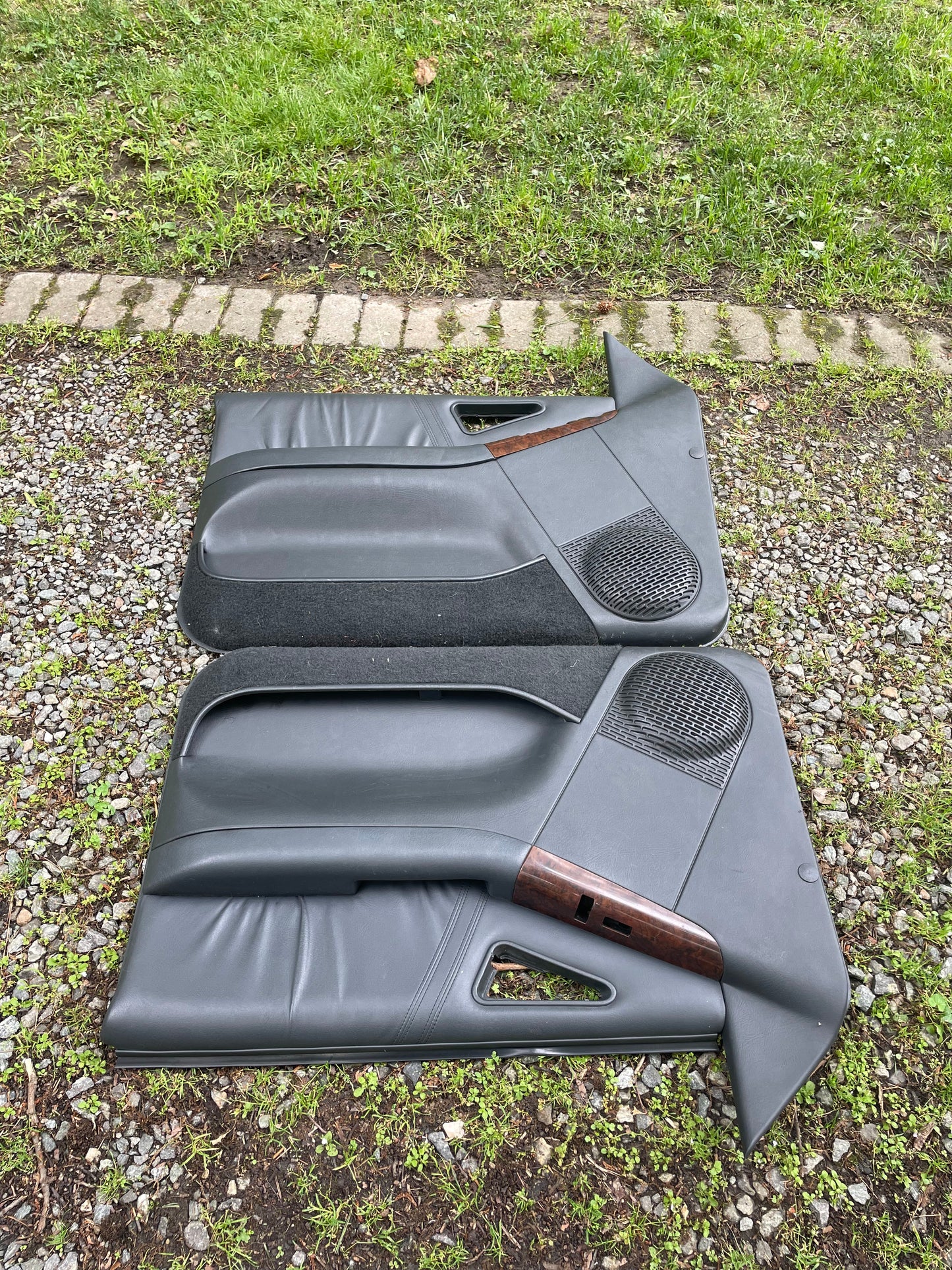 A clean OEM pair of dark gray front power door panels with woodgrain trim by General Motors, for 1994–1997 Chevy S10, GMC Sonoma, and more, are shown side by side outdoors on gravel and grass, ready for display or sale.