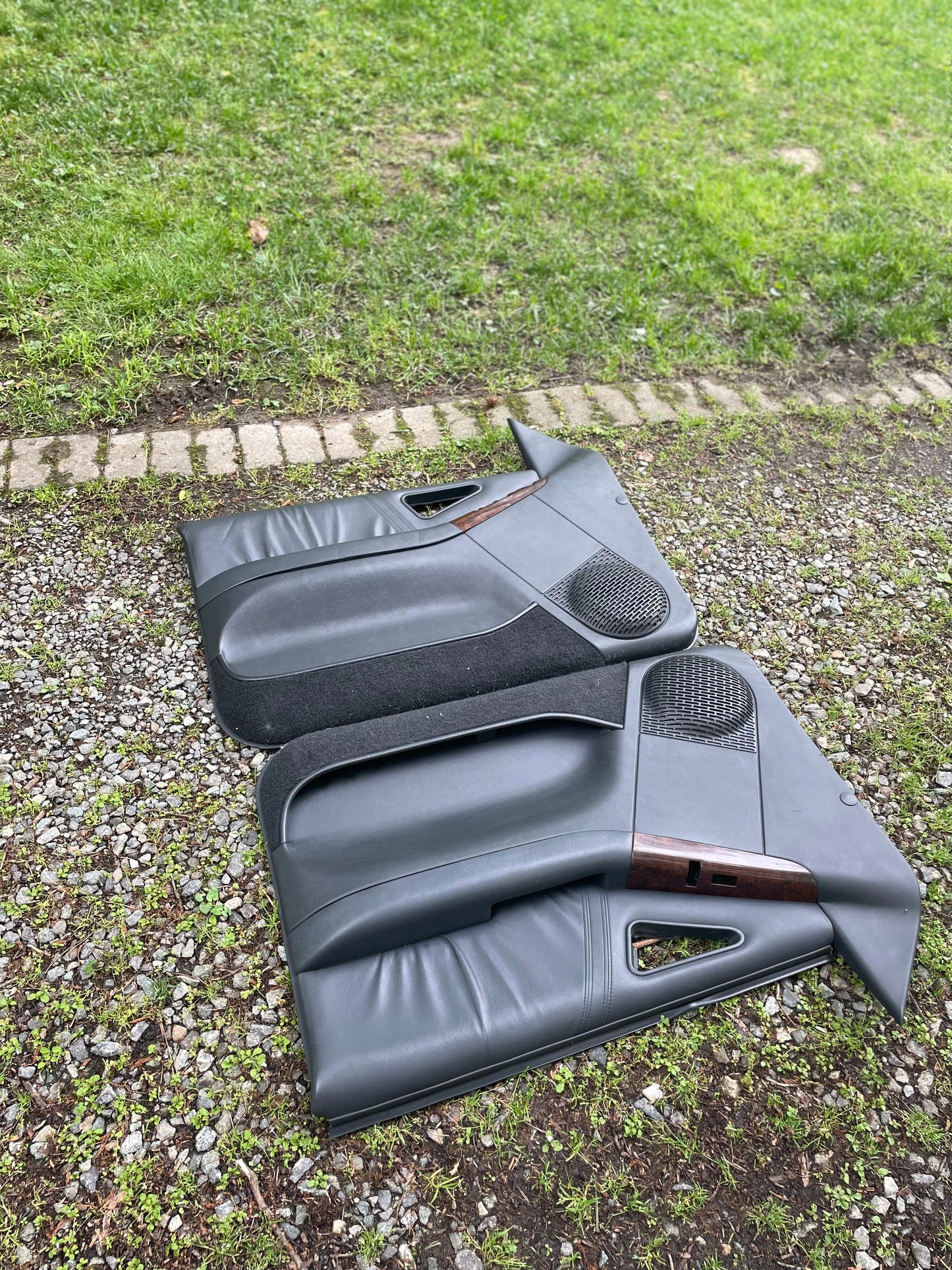 A General Motors OEM pair of front power door panels in dark gray with woodgrain for 1994-1997 Chevy S10, GMC Sonoma, and more are lying outdoors on gravel and grass near a stone path.