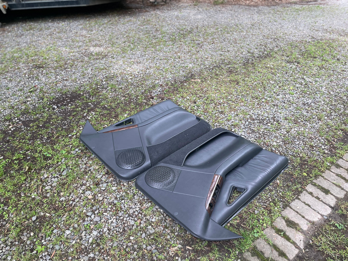 A pair of General Motors OEM front power door panels in dark gray with woodgrain, for 1994-1997 Chevy S10/GMC Sonoma, are lying on a gravel driveway next to a grassy area.