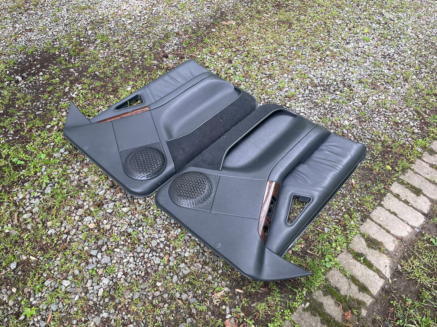 A pair of OEM General Motors dark gray front power door panels with woodgrain trim for 1994-1997 Chevy S10 or GMC Sonoma are lying outdoors on grass, gravel, and paving stones.