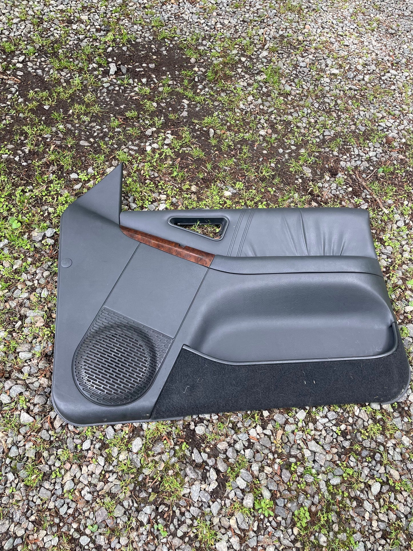 A General Motors OEM pair of front power door panels in dark gray with woodgrain for 1994-1997 Chevy S10, GMC Sonoma, and more lies outdoors on gravel and grass.