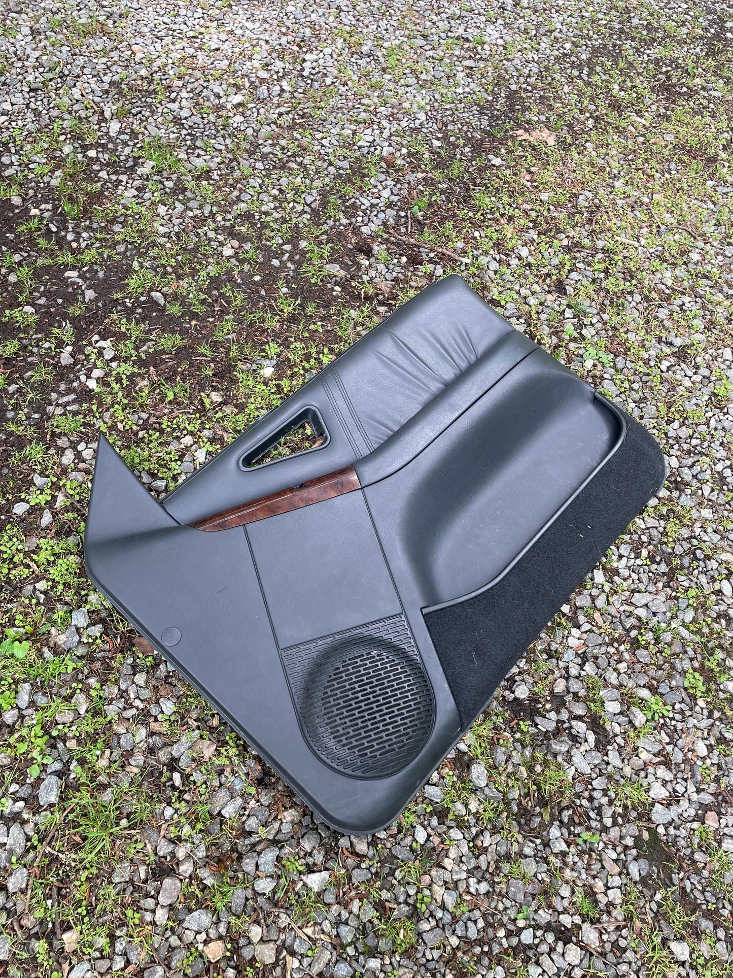 A pair of dark gray OEM front power door panels with woodgrain trim for 1994-1997 Chevy S10/GMC Sonoma by General Motors is shown resting outdoors on gravel and grass.