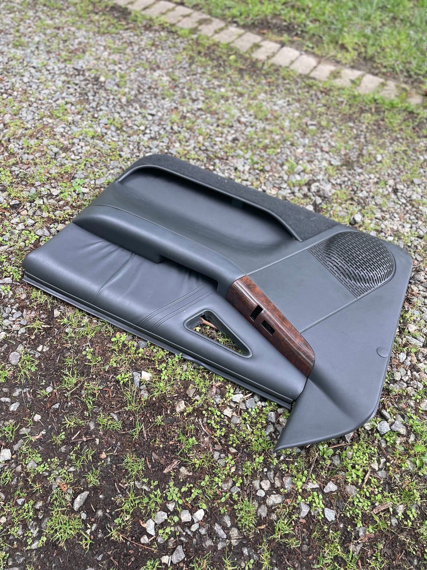 A General Motors OEM pair of front power door panels in dark gray with woodgrain, made for 1994-1997 Chevy S10 and GMC Sonoma, is shown outdoors on gravel and grass.