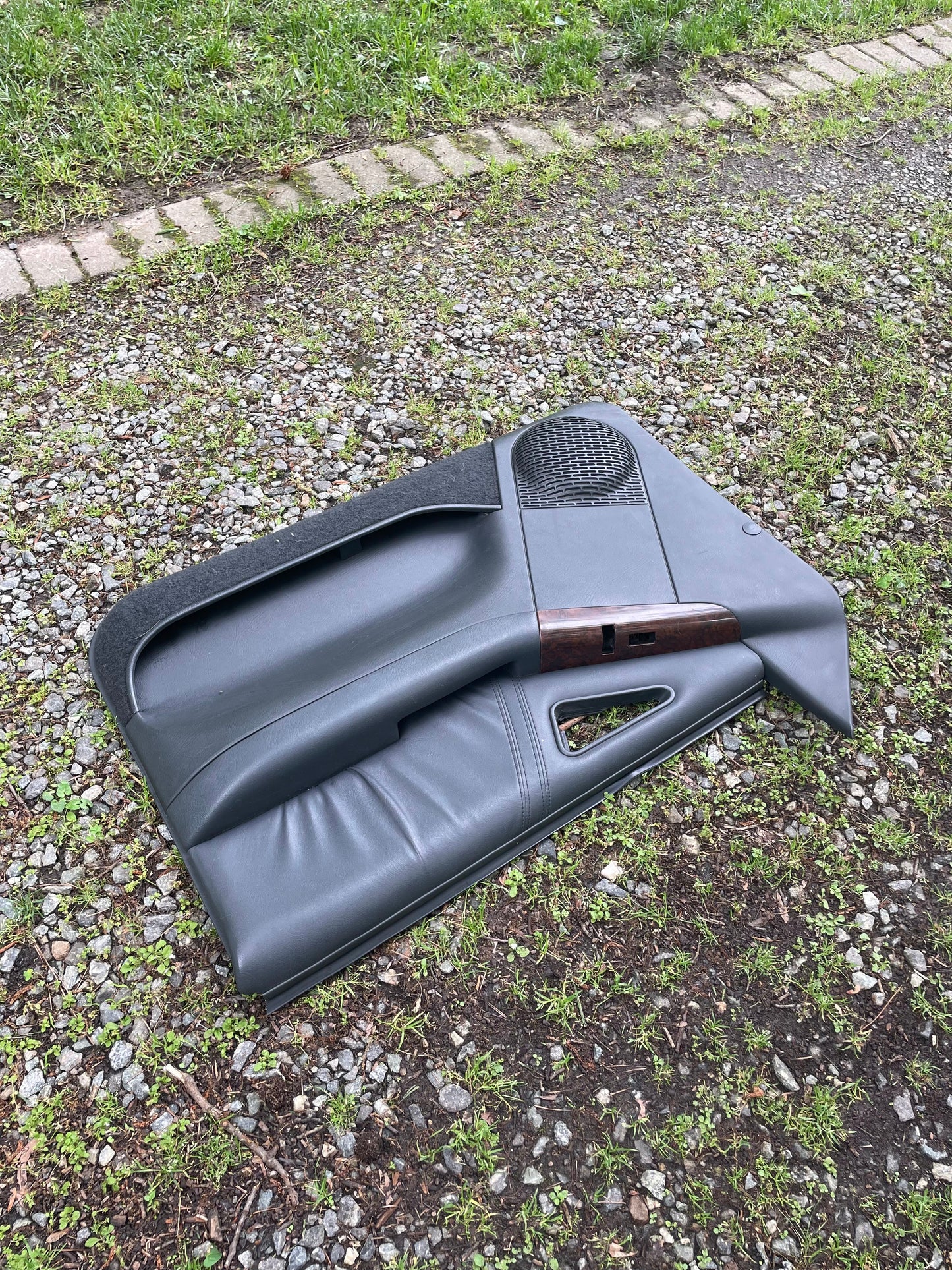A pair of OEM General Motors front power door panels in dark gray with woodgrain for 1994-1997 Chevy S10 and GMC Sonoma lies outdoors on gravel and grass near a brick border.