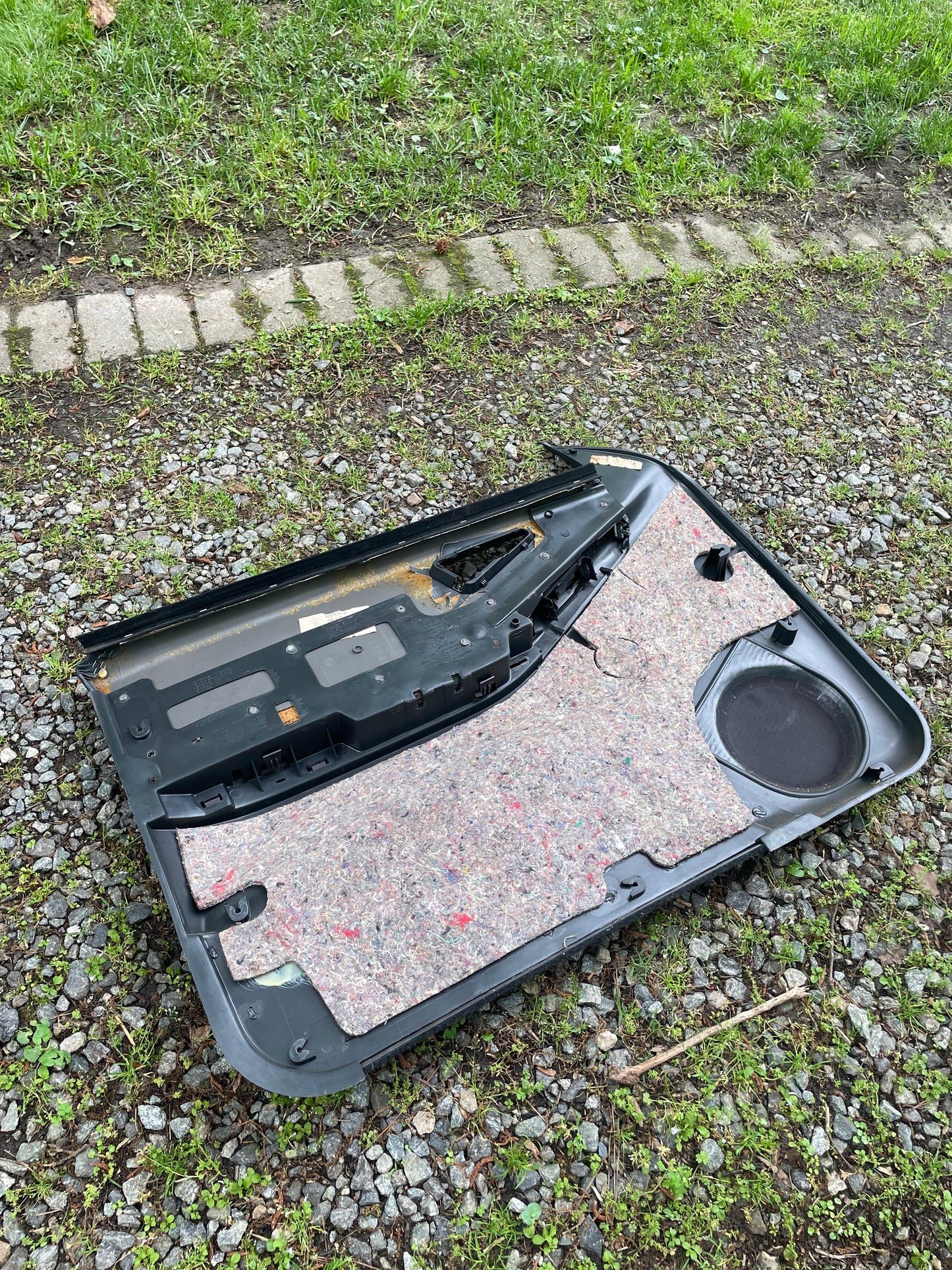 A detached OEM dark gray power door panel with woodgrain from a 1994-1997 Chevy S10 or GMC Sonoma by General Motors lies on gravel and grass, inner lining and speaker area exposed near gray bricks.