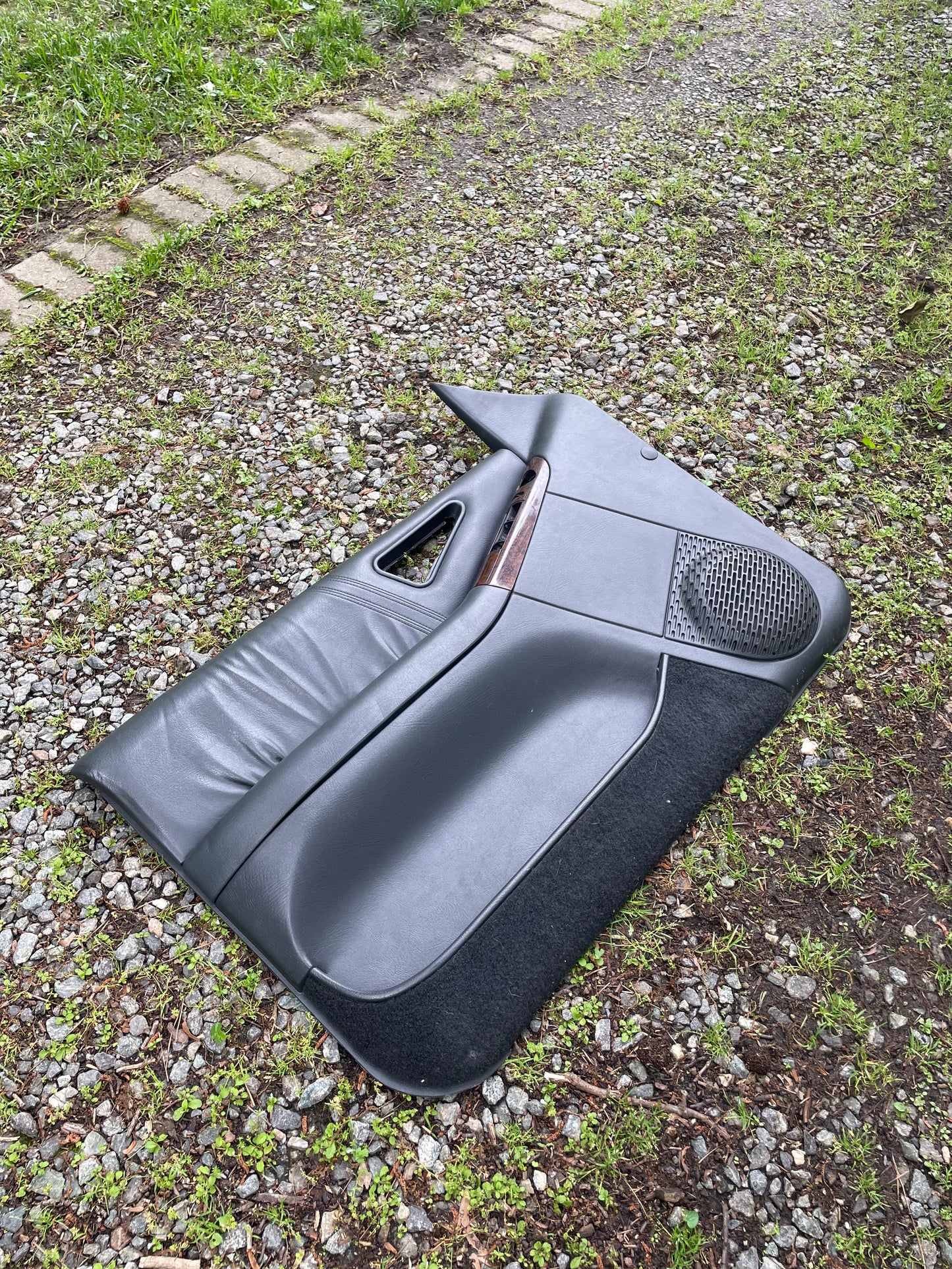 A pair of dark gray OEM General Motors front power door panels with woodgrain trim for 1994-1997 Chevy S10/GMC Sonoma rests on a gravel path, with grass in the background.