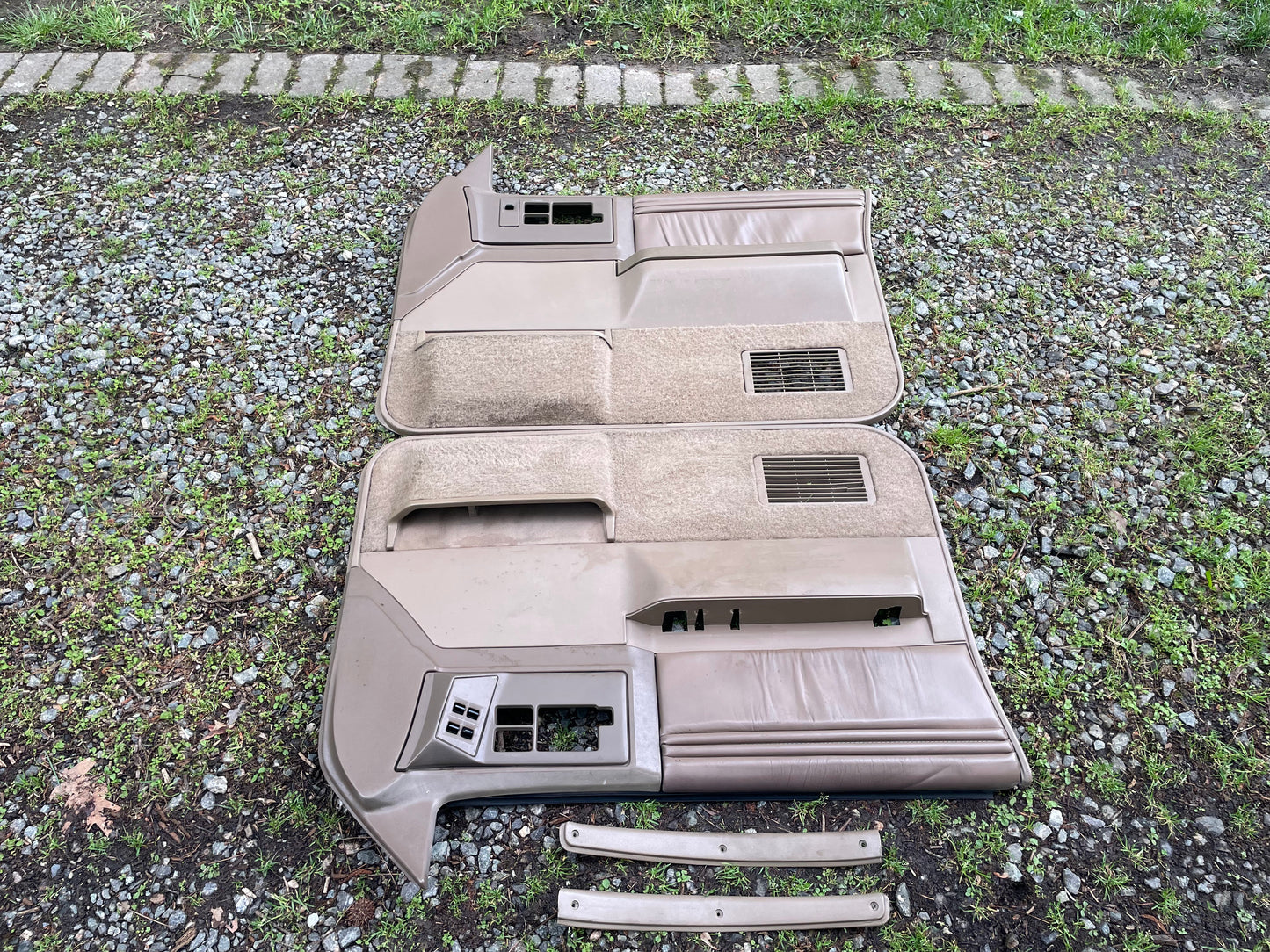 A General Motors OEM pair of beige leather front power door panels for 1986-1993 Chevy S10 & GMC S15, with control cutouts and matching trim pieces, are shown laid on gravel and grass.