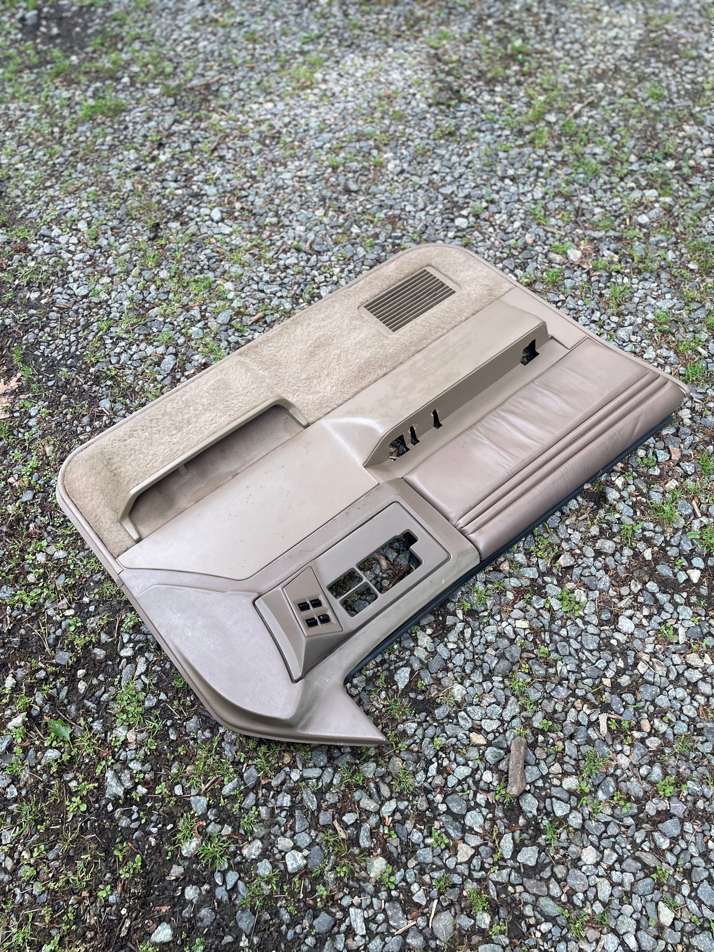 A General Motors OEM pair of beige leather front power door panels for 1986-1993 Chevy S10 and GMC S15 rests on gravel and grass, showing empty slots for switches or handles.