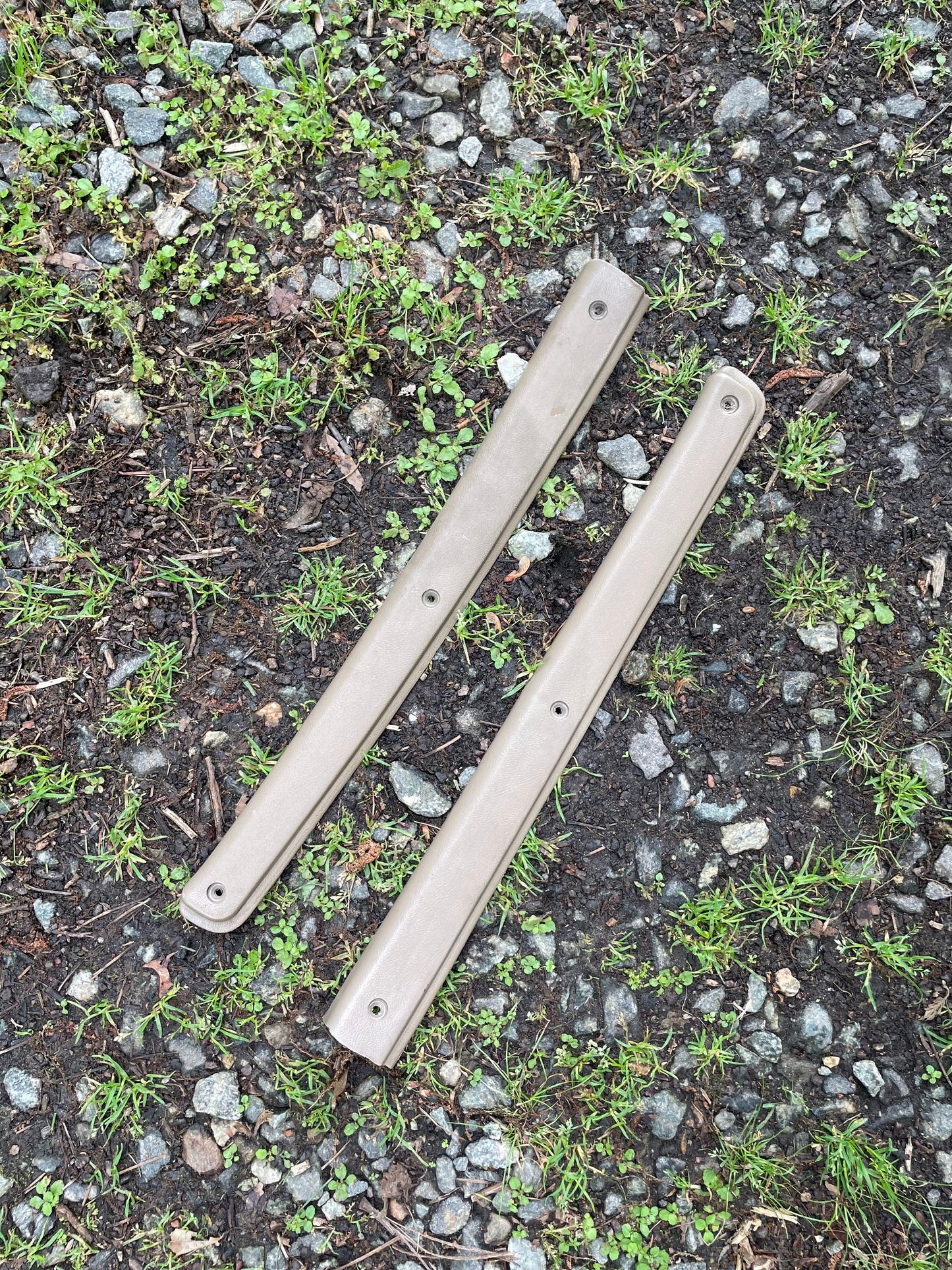 A pair of beige OEM leather front power door panels by General Motors for 1986-1993 Chevy S10 and GMC S15, lay parallel on a gravel and grass surface outdoors.
