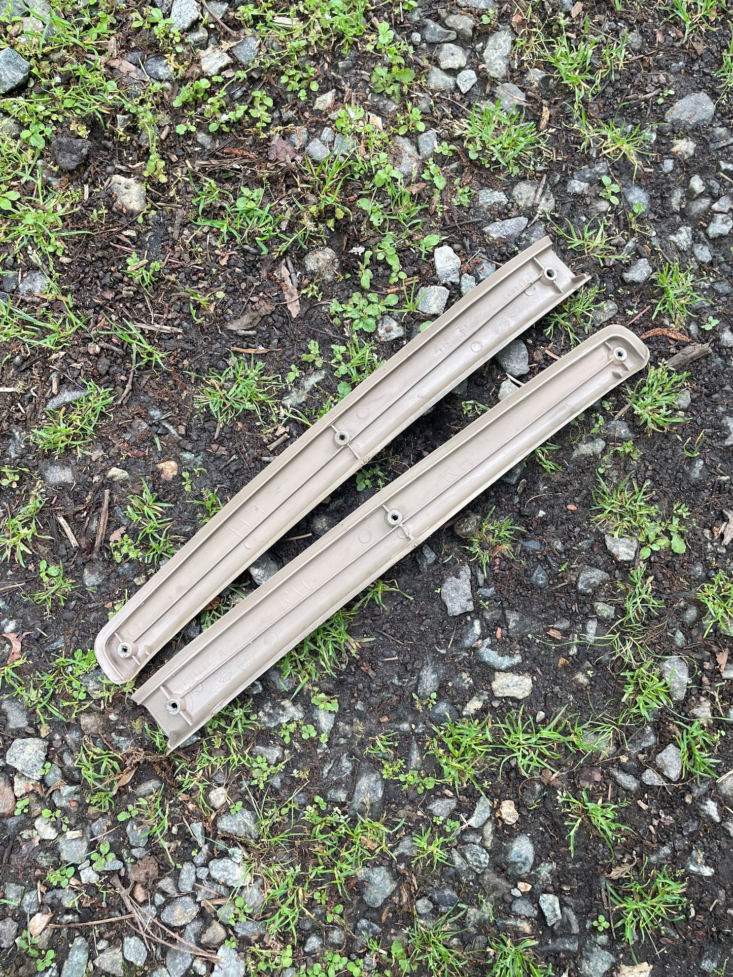 An OEM pair of leather front power door panels in beige for 1986-1993 Chevy S10/GMC S15 by General Motors rests parallel on gravel, surrounded by grass and small plants.