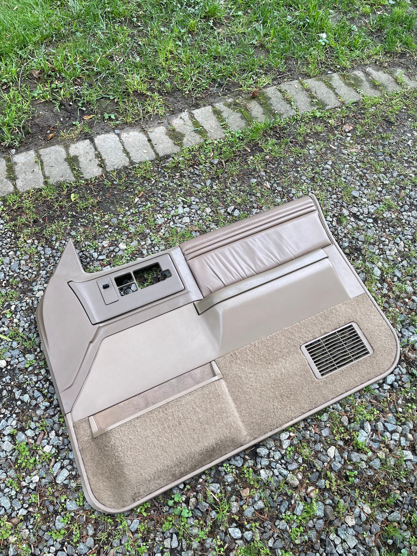 A General Motors OEM pair of beige leather front power door panels for 1986-1993 Chevy S10/GMC S15 lies on gravel beside grass bordered by paving stones, including empty slots for power window switches and vents.
