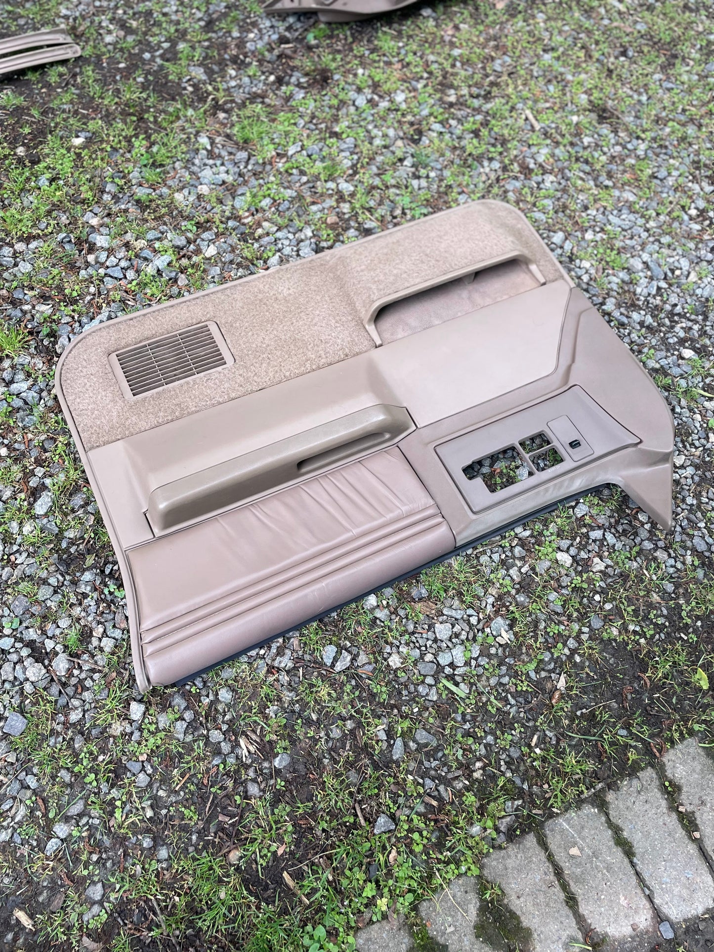 An OEM pair of beige leather front power door panels by General Motors for 1986-1993 Chevy S10 and GMC S15 is shown on gravel and grass, featuring a handle, armrest, vent, and power window switch openings.