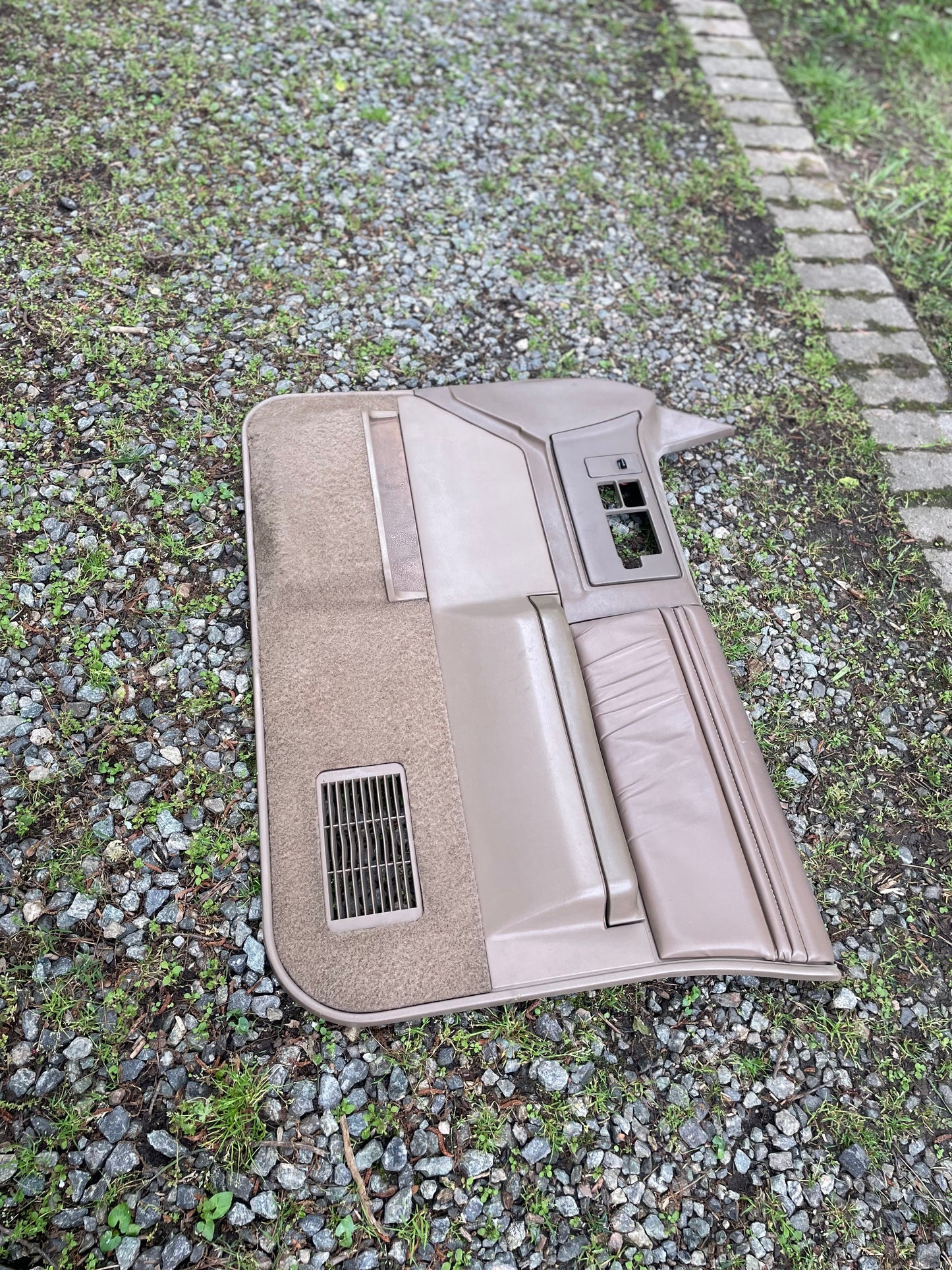 An OEM pair of beige leather front power door panels by General Motors for 1986-1993 Chevy S10, GMC S15, and more lies on gravel and grass next to a row of concrete pavers.