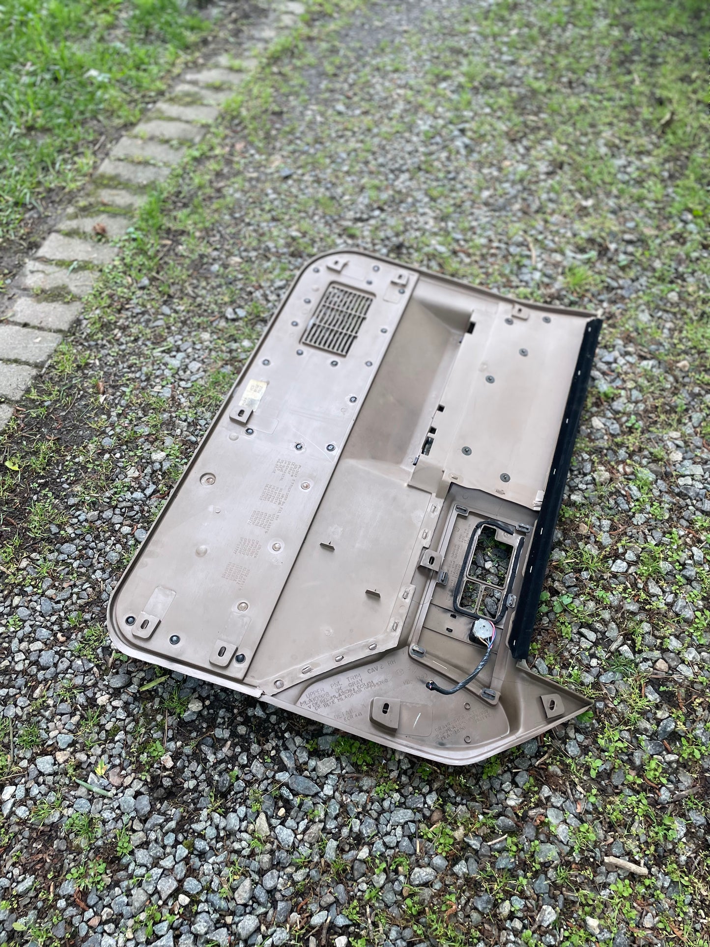 A broken beige plastic component, likely from a General Motors OEM Pair of Leather Front Power Door Panels (1986-1993 Chevy S10/GMC S15), lies face down outdoors on gravel and grass, exposing internal wiring and circuit board.