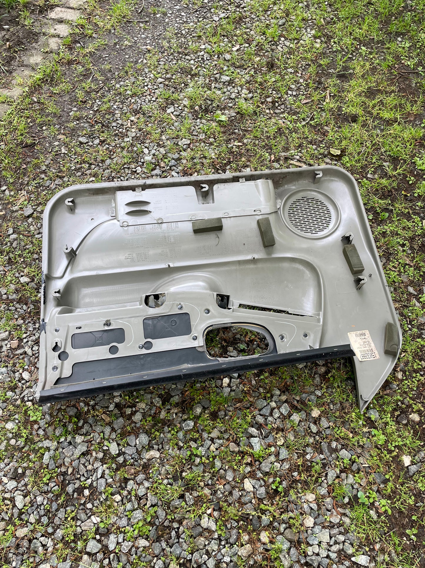 A General Motors OEM front power door panel in light gray for 1998-2005 Chevy S10/GMC Sonoma rests outdoors on gravel and grass, showing its inner structure and speaker cover.