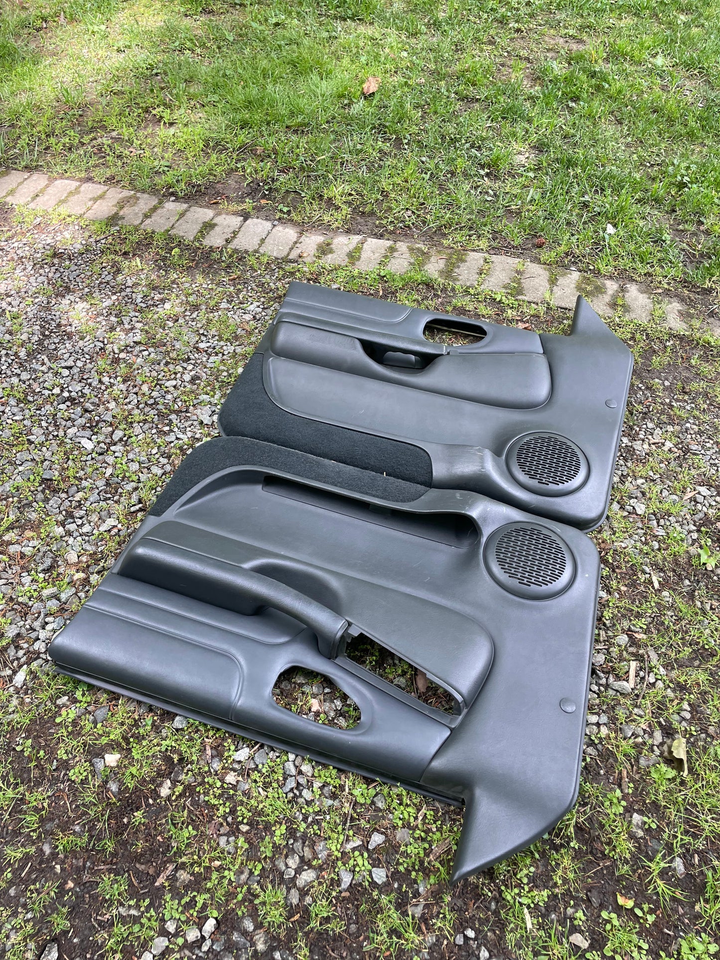 A pair of OEM General Motors front power leather door panels in dark gray for 1998-2005 Chevy S10/GMC Sonoma are lying outdoors on grass and gravel, parallel to each other, with paving stones visible in the background.