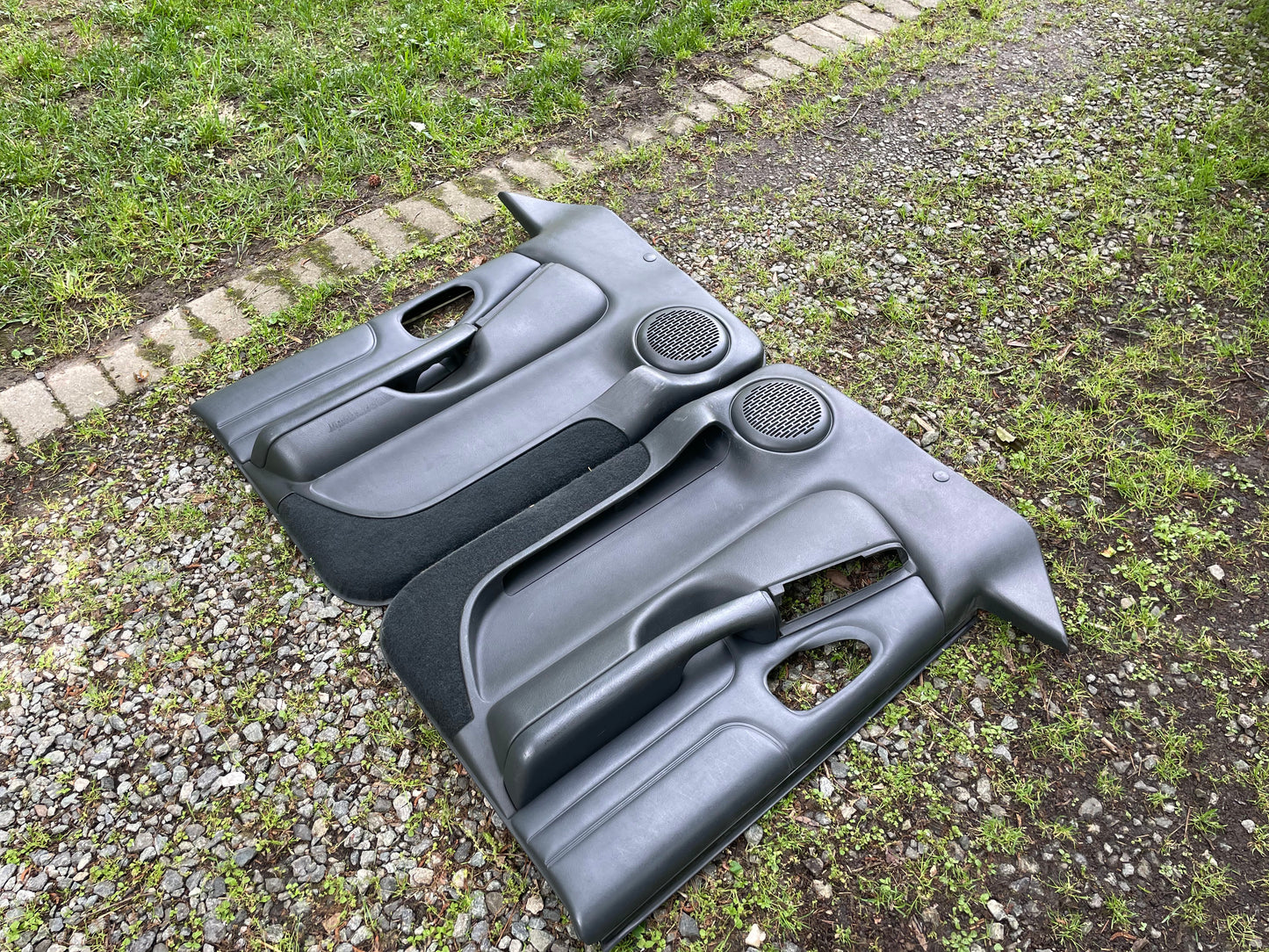 A pair of OEM General Motors front power leather door panels in dark gray for 1998-2005 Chevy S10/GMC Sonoma are side by side on gravel and grass by a stone-edged path.