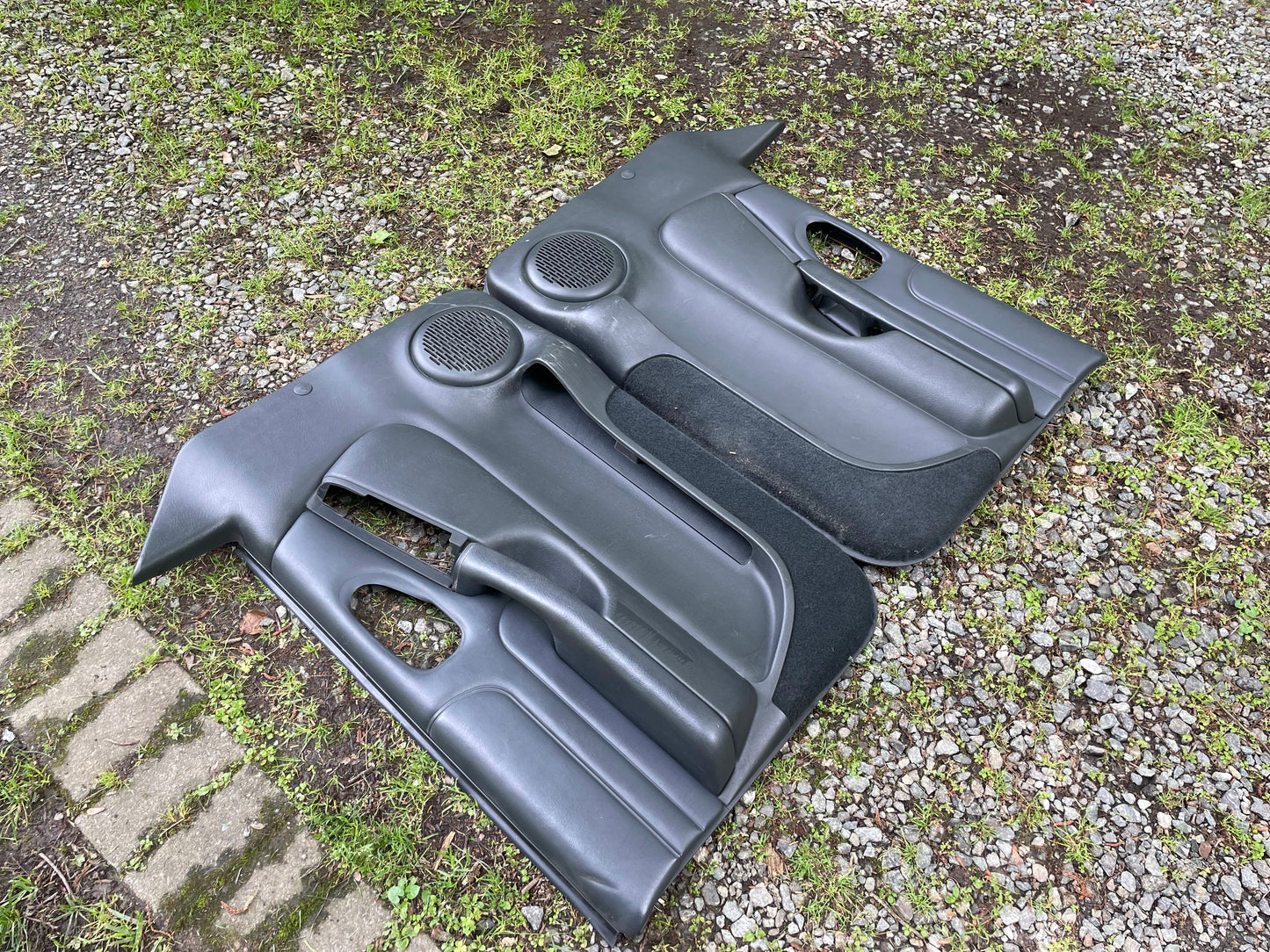 A pair of OEM General Motors front power leather door panels in dark gray for 1998-2005 Chevy S10/GMC Sonoma are shown outdoors on gravel and grass.