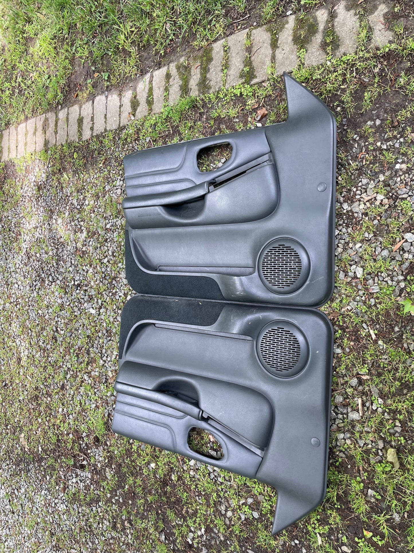 An OEM pair of front power leather door panels in dark gray for 1998-2005 Chevy S10 and GMC Sonoma by General Motors are lying side by side outdoors on grass and gravel, with speaker grills and armrests visible; a stone path runs above them.