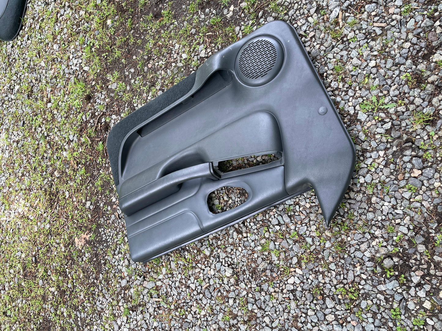 A pair of General Motors OEM front power leather door panels in dark gray for 1998-2005 Chevy S10/GMC Sonoma, featuring speaker grilles and handles, rest on gravel and grass outdoors.