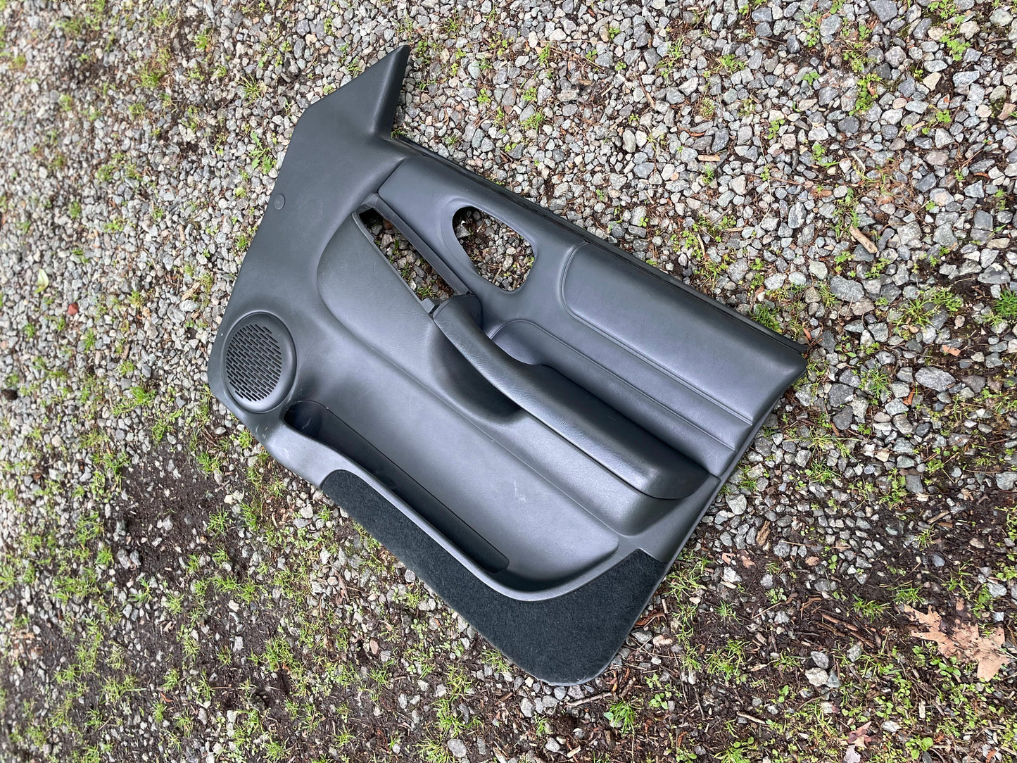 An OEM pair of dark gray leather front door panels for 1998-2005 Chevy S10/GMC Sonoma by General Motors, designed for power windows and locks, rests on gravel with grass and soil patches.
