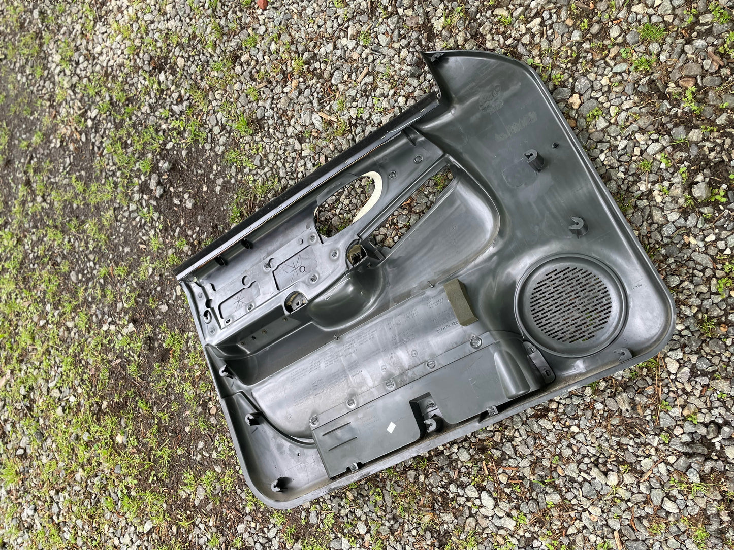 A pair of OEM General Motors dark gray front power leather door panels for 1998-2005 Chevy S10/GMC Sonoma, featuring speaker grilles and handle openings, are shown lying on gravel with scattered green moss.