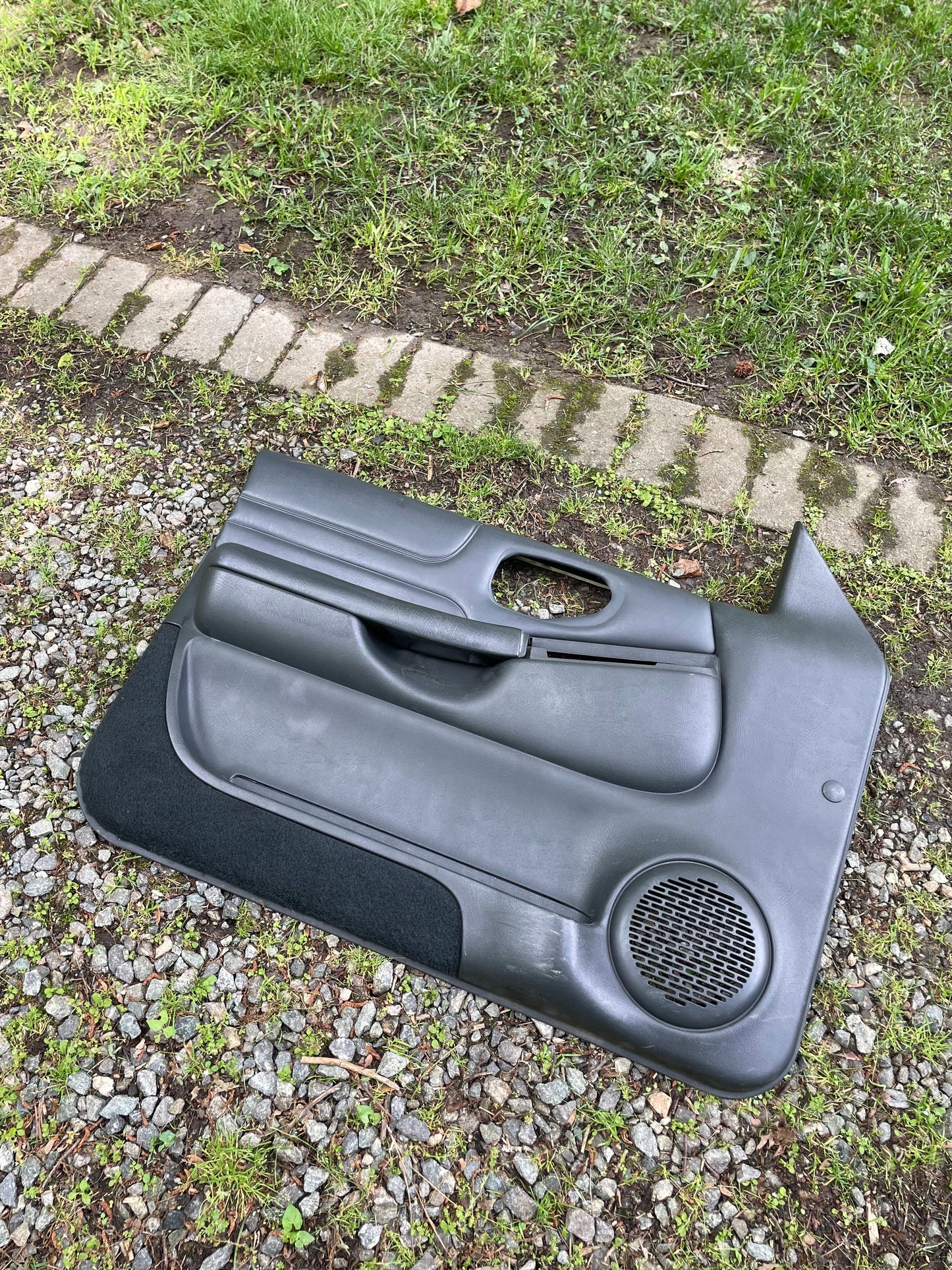 A pair of OEM General Motors dark gray leather front power door panels for 1998-2005 Chevy S10/GMC Sonoma, with built-in speakers, power windows and locks, rests on gravel near grass and a brick border.