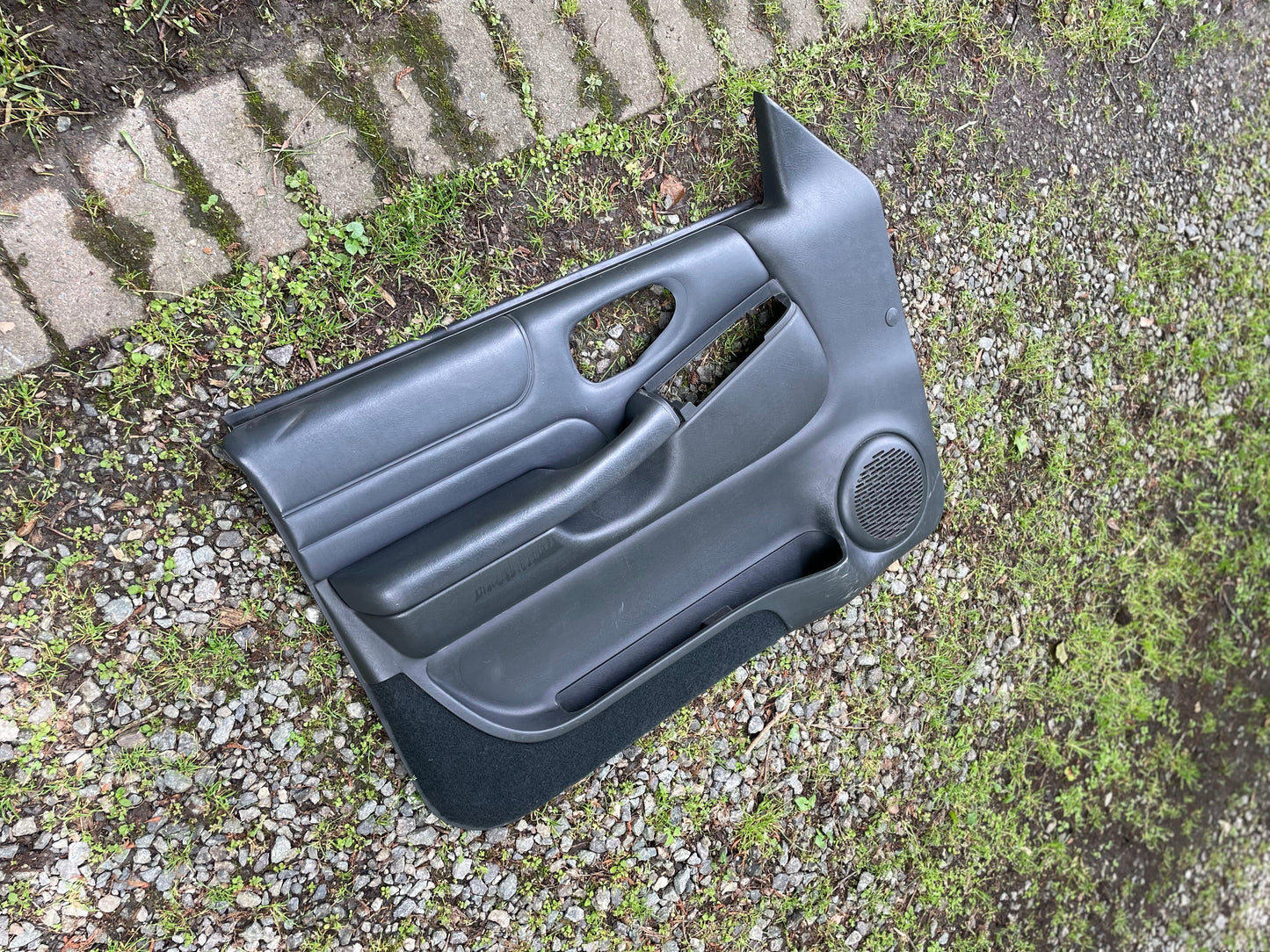A General Motors OEM pair of front power leather door panels in dark gray for 1998-2005 Chevy S10 and GMC Sonoma lies on the ground among small rocks, patches of grass, and a stone walkway.