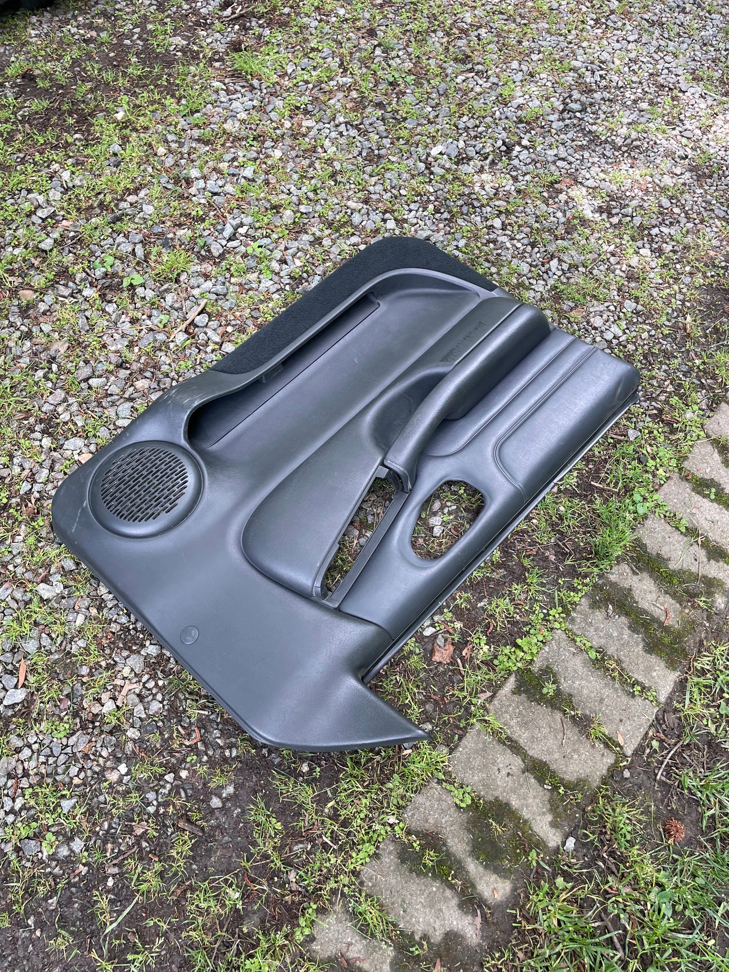 An OEM pair of front power leather door panels in dark gray for 1998-2005 Chevy S10 and GMC Sonoma by General Motors rests outdoors on grass and gravel near concrete stepping stones, with visible speaker grilles.
