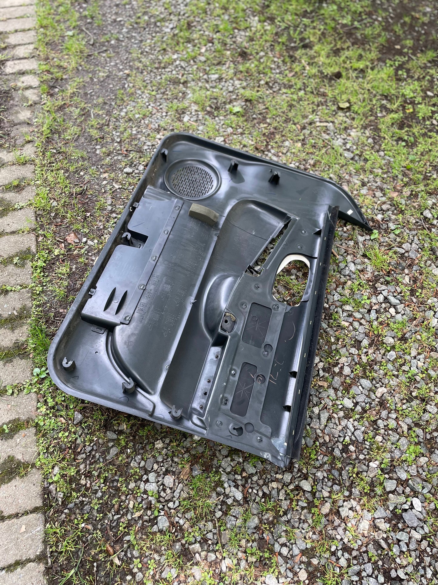 An OEM pair of front power leather door panels in dark gray by General Motors for 1998-2005 Chevy S10 and GMC Sonoma lies face-up on gravel and grass, with concrete pavers visible on the left side.