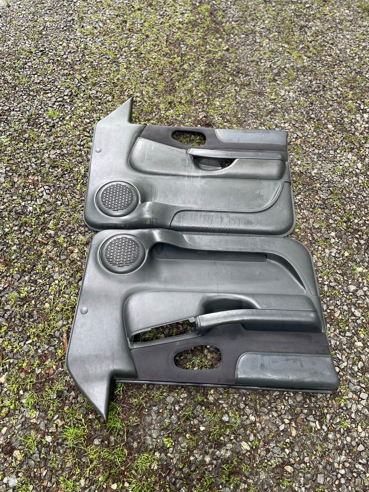 OEM Pair of Front Power Door Panels in Dark Gray for 1998-2005 Chevy S10 and GMC Sonoma by General Motors with speaker grilles and handle cutouts. Excellent upgrade for your interior, pictured side by side outdoors.
