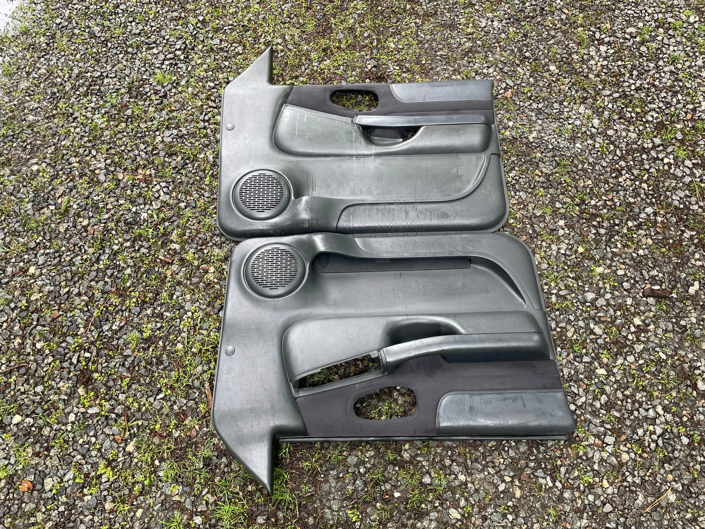 A pair of General Motors OEM front power door panels in dark gray for 1998-2005 Chevy S10/GMC Sonoma lie side by side on gravel and grass, showing wear and featuring built-in speaker areas plus openings for handles and controls.