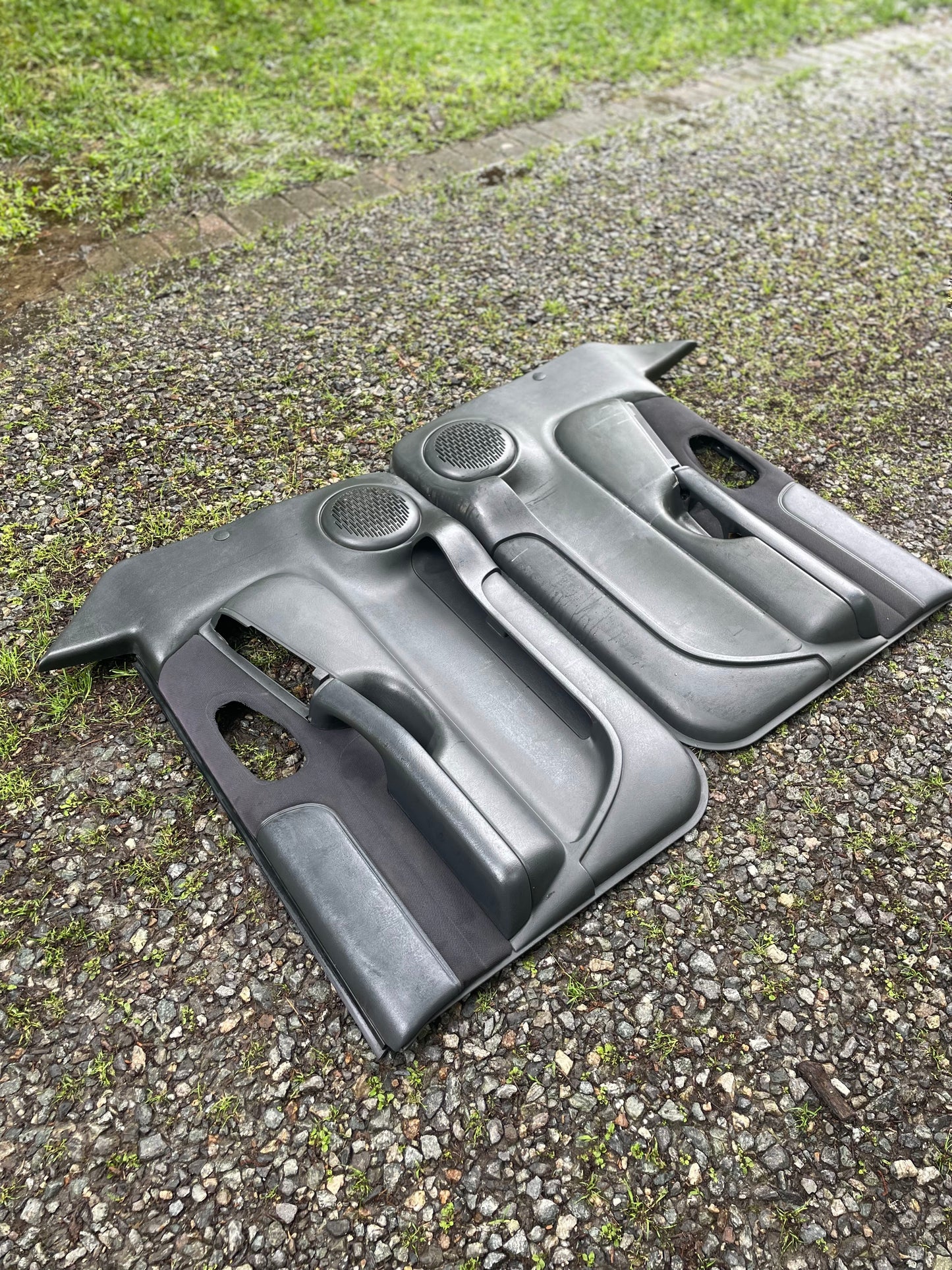 A pair of OEM General Motors front power door panels in dark gray for 1998-2005 Chevy S10, GMC Sonoma and more, are shown on gravel with grass—ideal originals for restoring your interior.