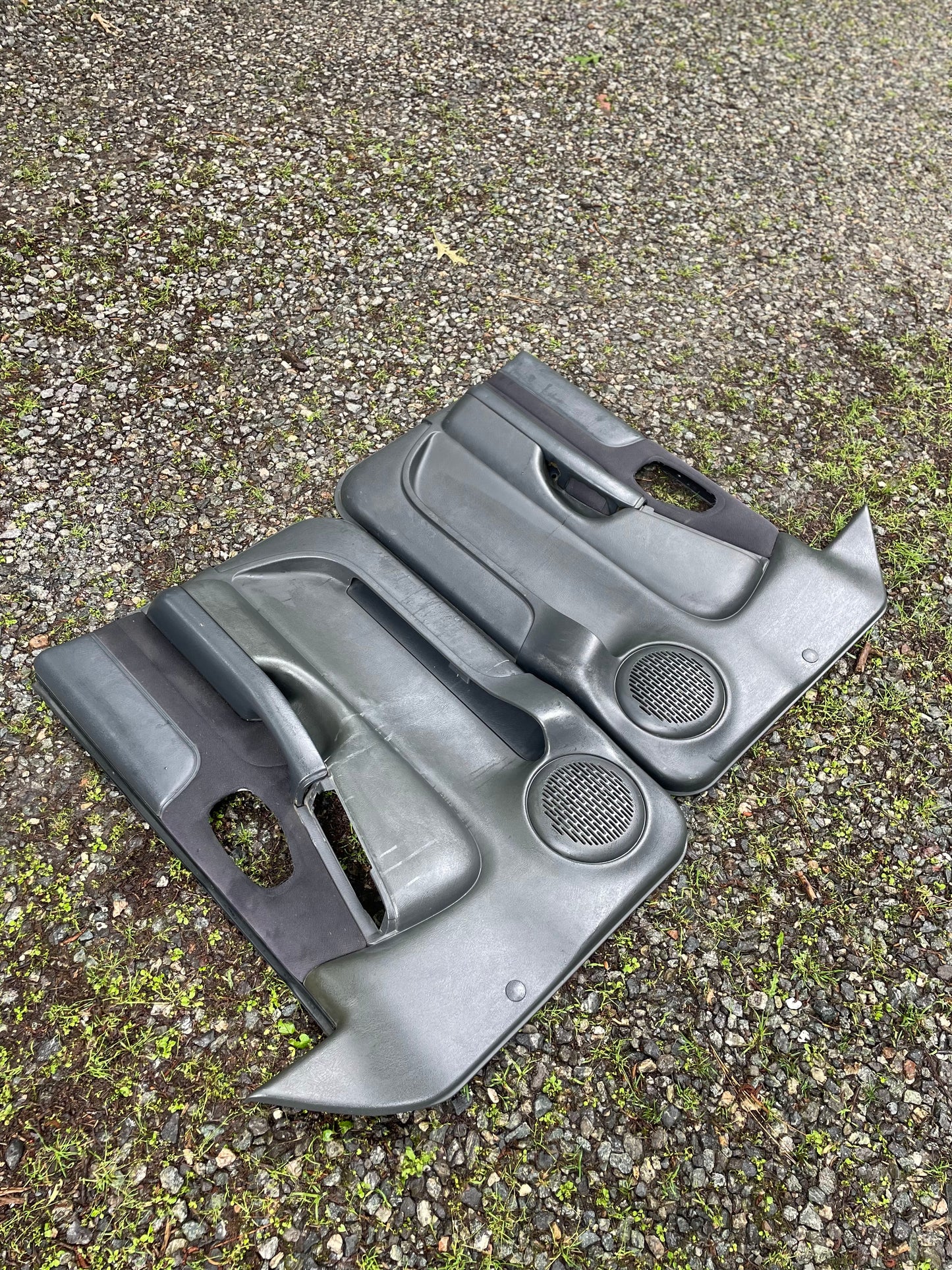 A pair of General Motors OEM dark gray front power door panels for 1998-2005 Chevy S10 and GMC Sonoma, showing visible wear and cutouts for window switches, are lying on gravel and grass.