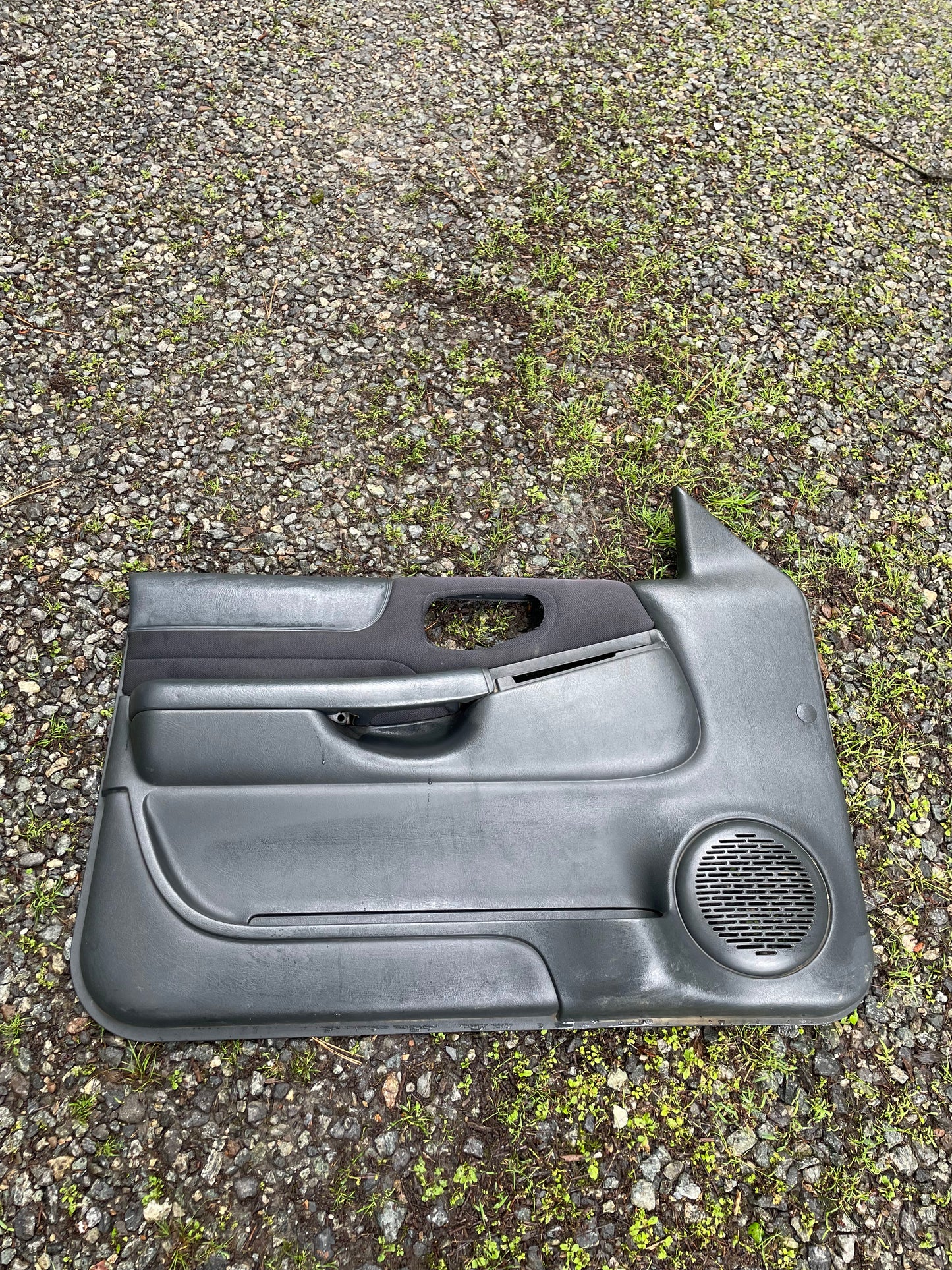 An OEM pair of General Motors front power door panels in dark gray for 1998-2005 Chevy S10, GMC Sonoma, and more is shown on a gravel surface with patches of green grass.