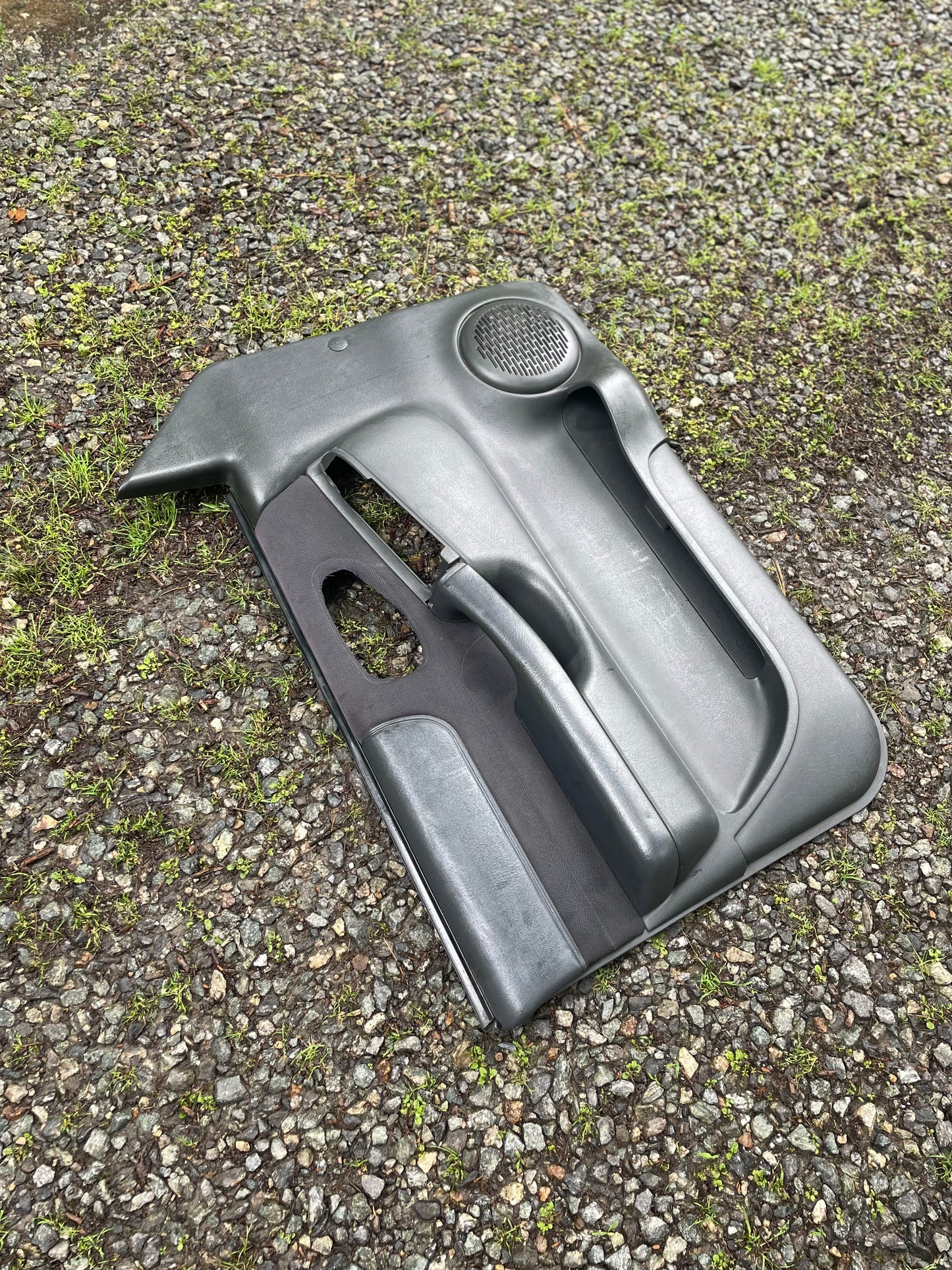 An OEM pair of General Motors front power door panels in dark gray for 1998-2005 Chevy S10 and GMC Sonoma is shown resting on a gravel surface with some green grass patches.