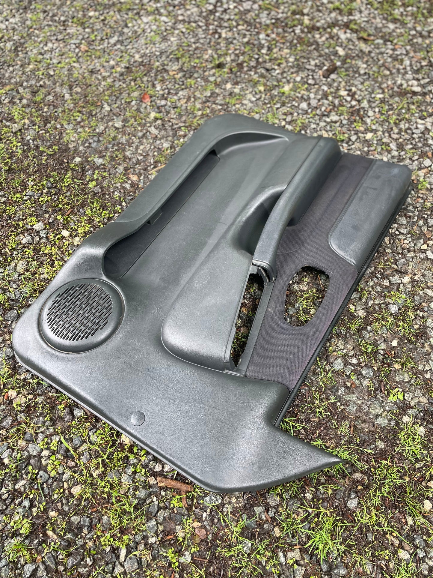 A pair of General Motors OEM dark gray front power door panels for 1998-2005 Chevy S10 and GMC Sonoma rests outdoors on gravel and green moss, featuring a circular speaker opening.