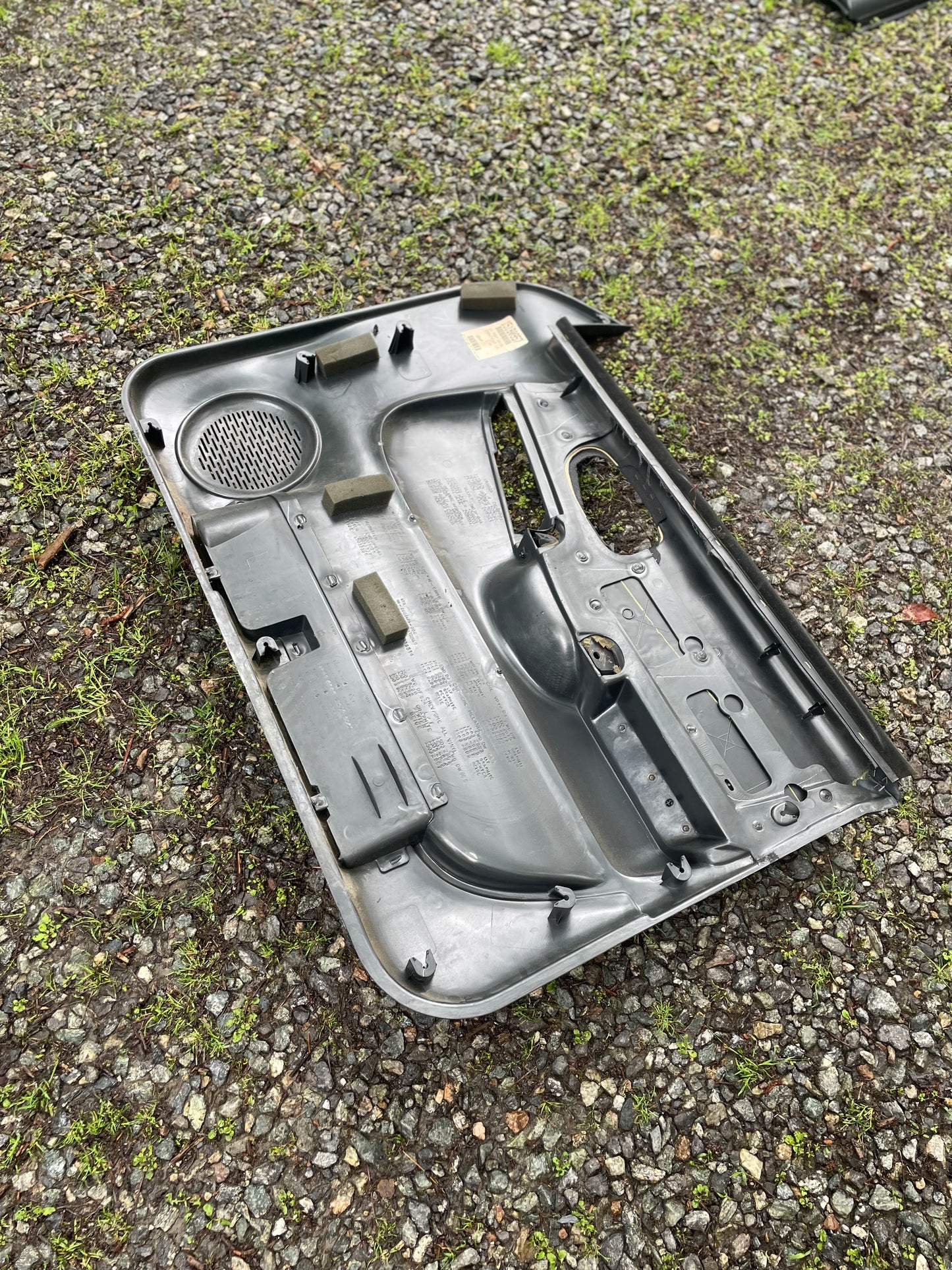 A General Motors OEM pair of dark gray front power door panels for 1998-2005 Chevy S10 and GMC Sonoma lies face down on gravel with green moss, showing molded shapes, a speaker grille, and attachment points on the underside.