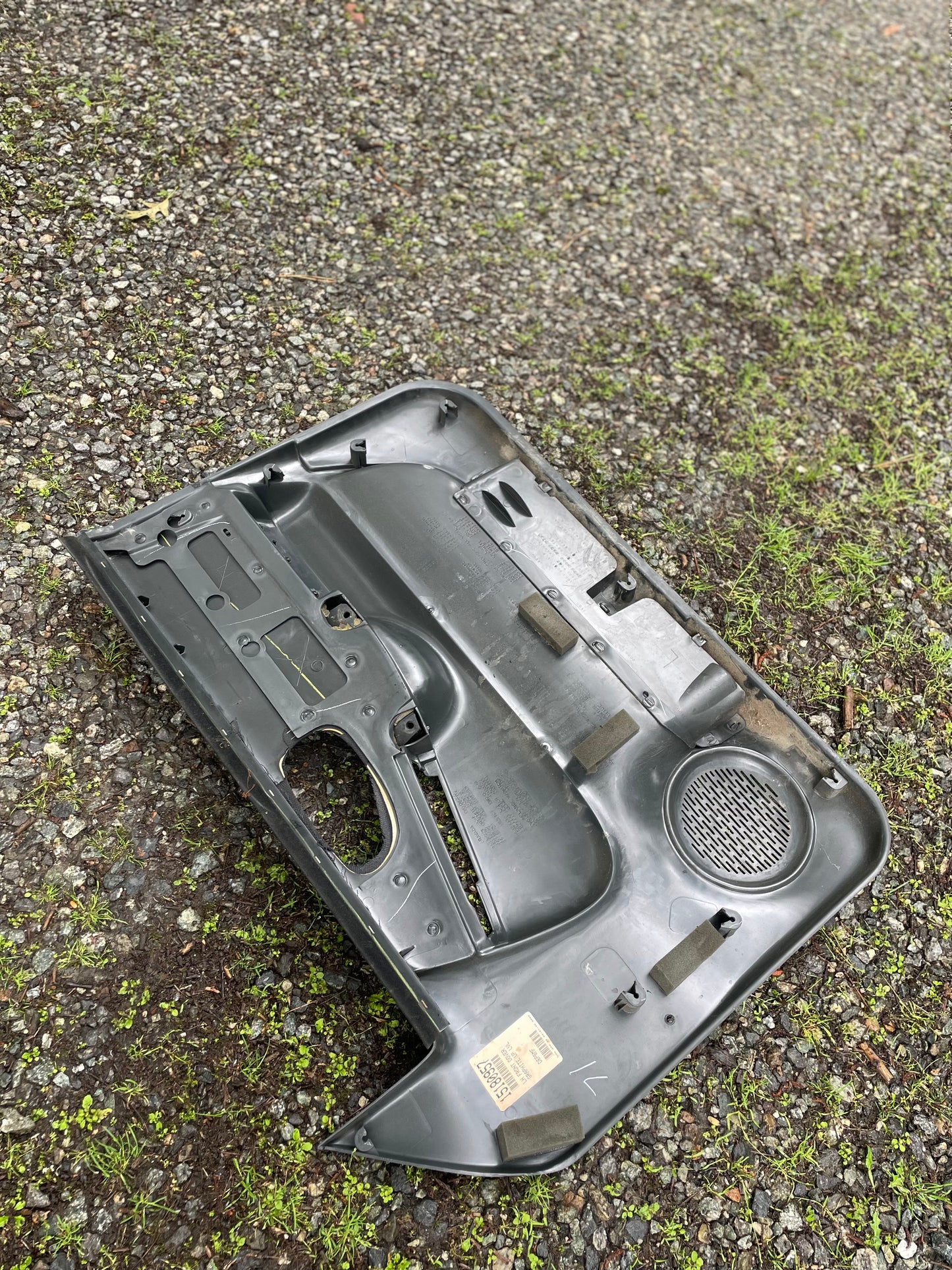 A pair of General Motors OEM dark gray plastic front power door panels for 1998-2005 Chevy S10/GMC Sonoma lies face down on gravel and grass, showing the speaker grille and handle cutouts.