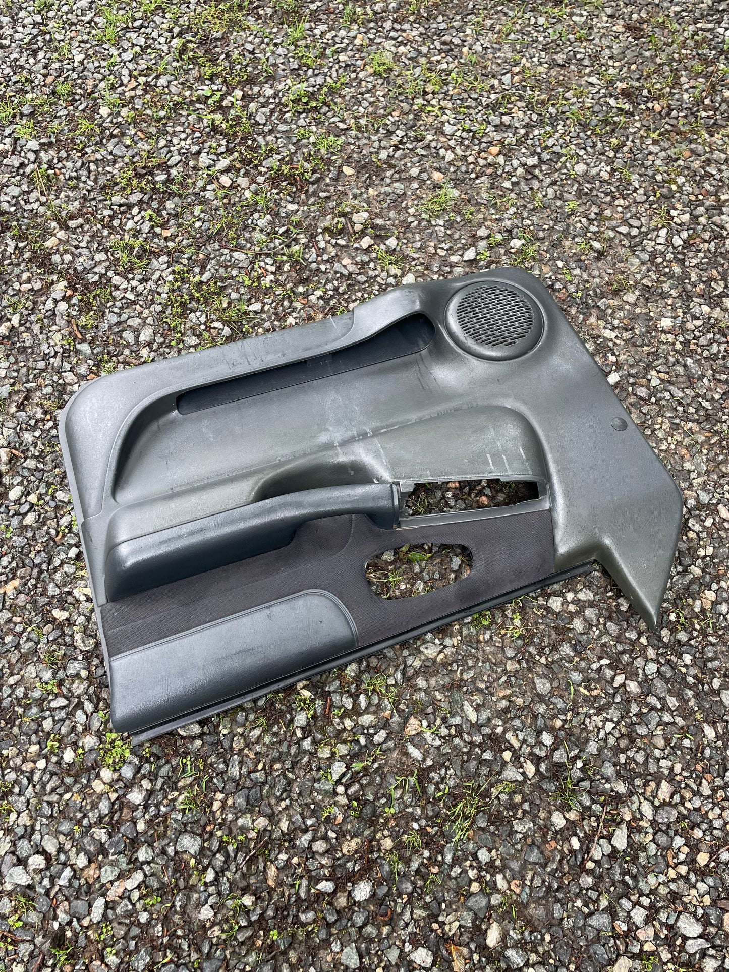 A General Motors OEM pair of front power door panels in dark gray for 1998-2005 Chevy S10 and GMC Sonoma is seen detached on a gravel surface, with integrated speaker and handle visible amid scattered grass.