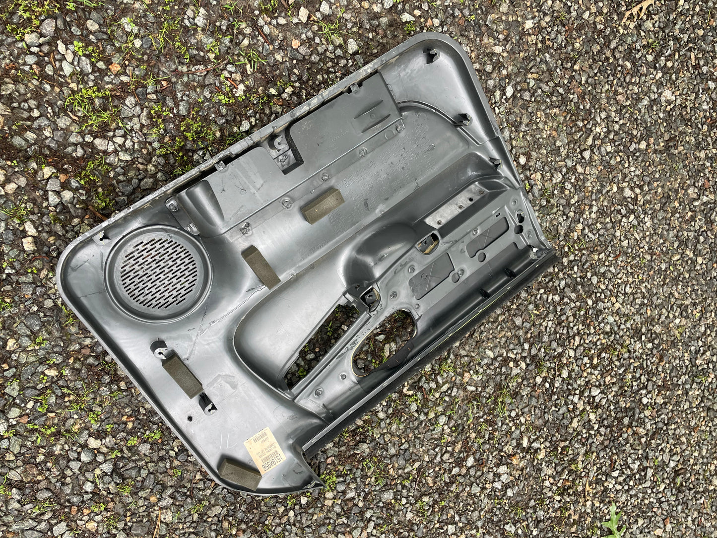 An OEM pair of General Motors dark gray front power door panels for 1998-2005 Chevy S10 and GMC Sonoma lies face down on gravel and mossy ground, featuring speaker grilles and compartments.