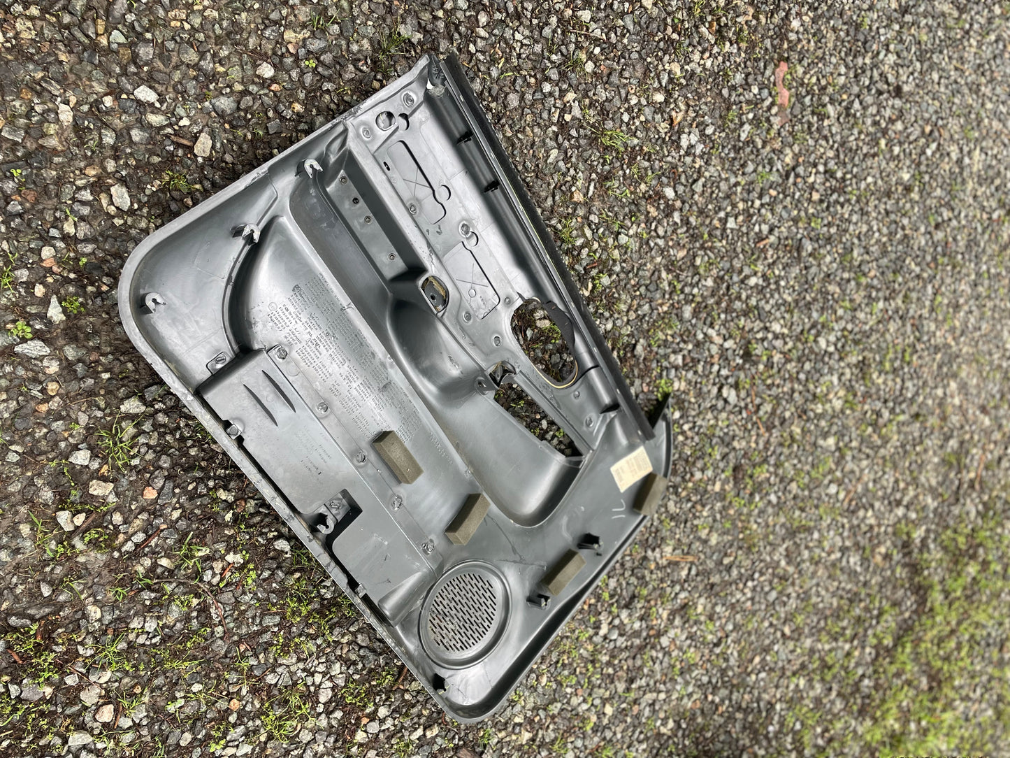 An OEM pair of front power door panels in dark gray by General Motors for 1998-2005 Chevy S10 or GMC Sonoma lies face down on gravel, with patches of green moss or grass visible among the stones.