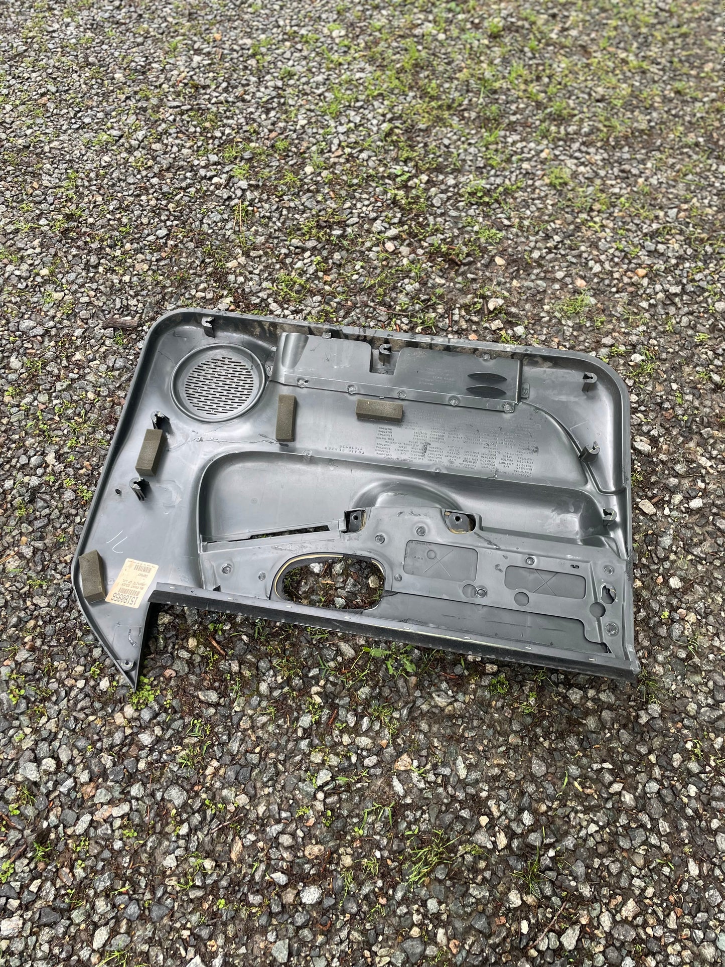 A General Motors OEM pair of front power door panels in dark gray for 1998-2005 Chevy S10 and GMC Sonoma is shown face down on gravel, displaying the interior side with visible cutouts and some grass poking through the stones.