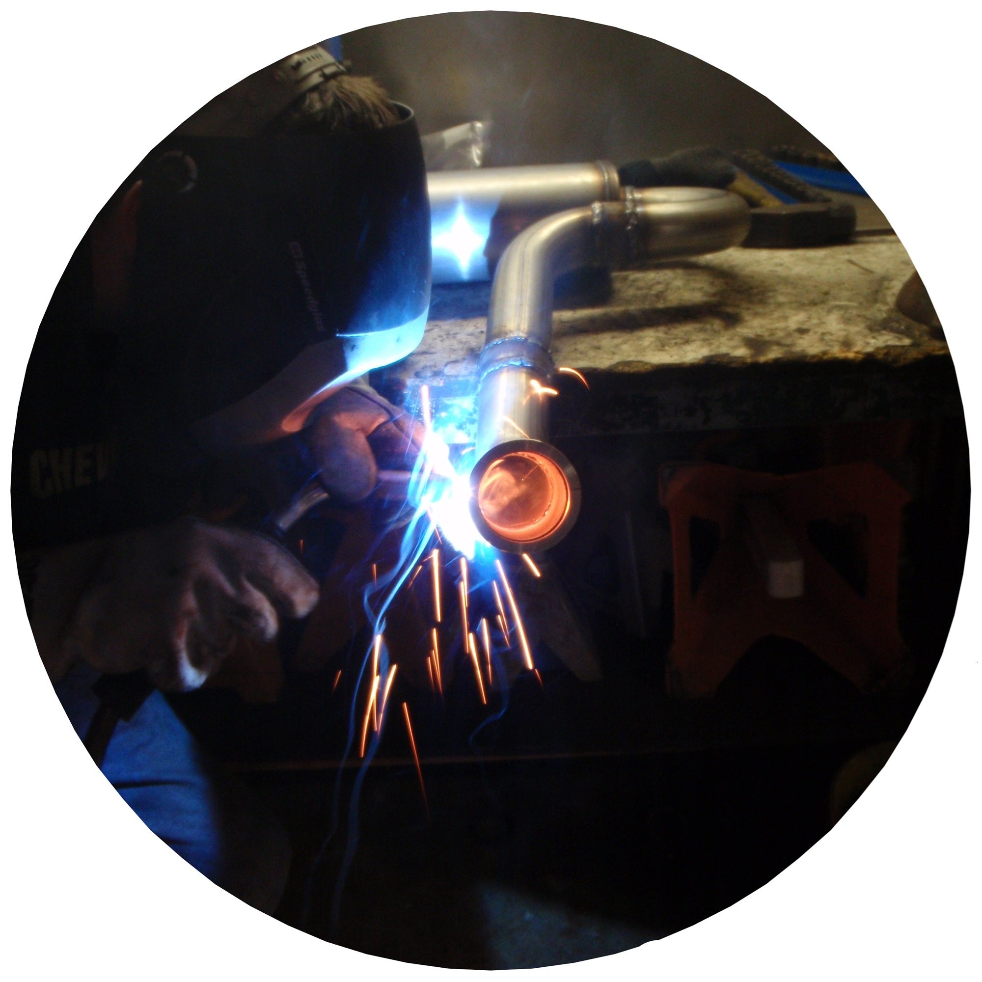 A person wearing protective gear is welding a metal pipe on a workbench, with bright sparks and blue light visible from the welding process.