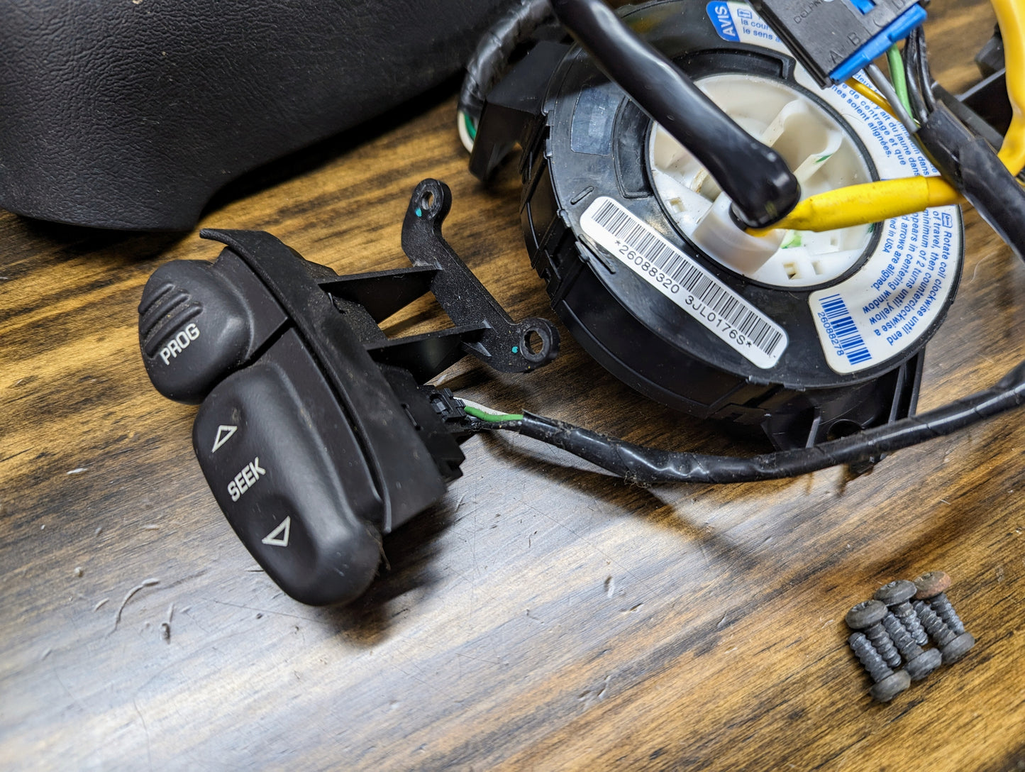 Close-up of General Motors KIT Steering Wheel Radio Controls & Clock Spring for 1995-2002 GM Full Size OBS/NBS Trucks, featuring black PROG and SEEK buttons with wires, screws, and car parts on a wooden surface.