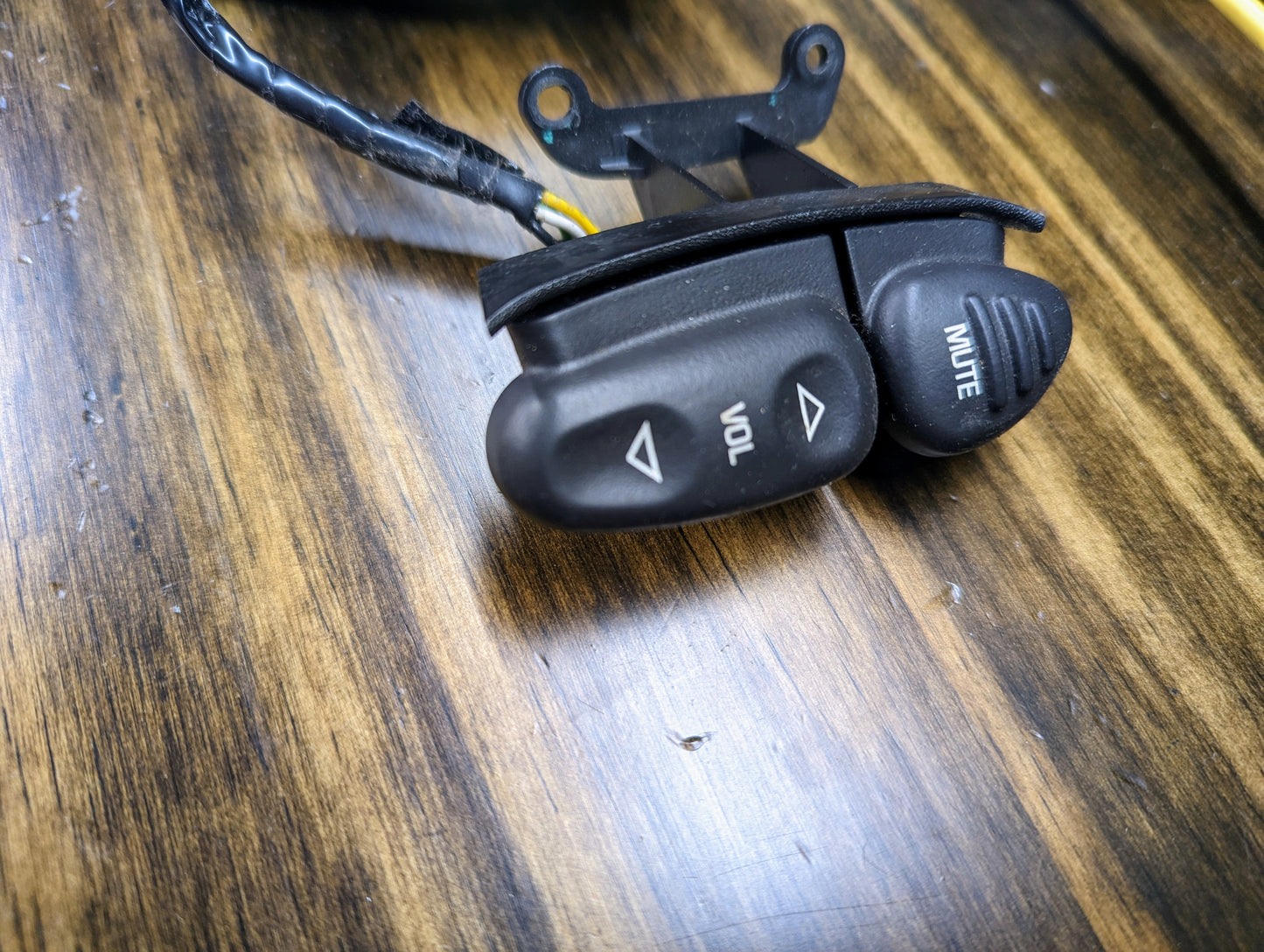 A close-up of a General Motors KIT Steering Wheel Radio Controls & Clock Spring for 1995-2002 GM Full Size OBS/NBS Trucks, featuring a black button labeled VOL with up/down arrows and MUTE, attached to a wire on a wood-grain surface.